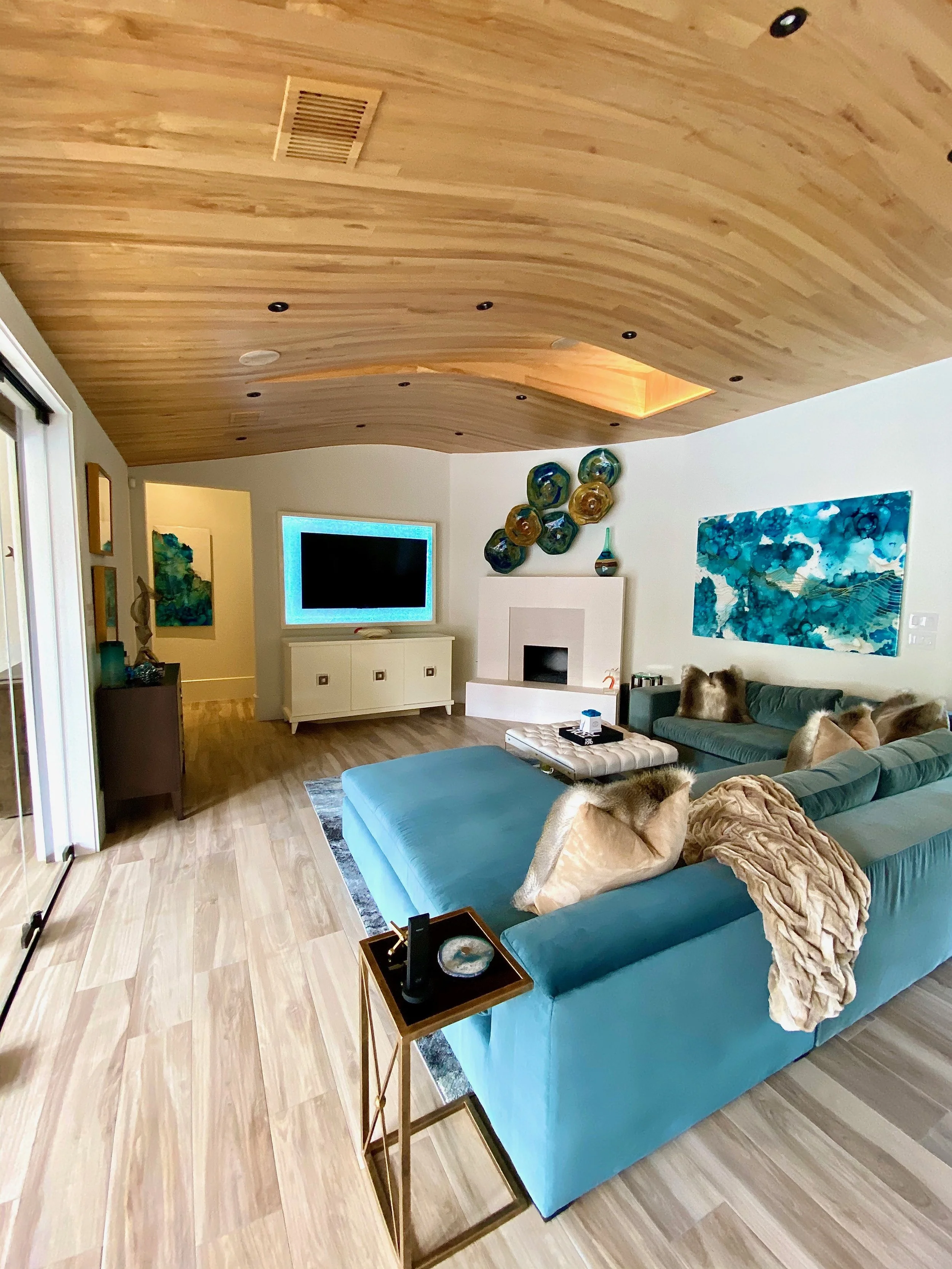 Modern Resort style luxury living room with curved natural wood ceiling, teal sectional sofa, custom millwork, custom fireplace, and abstract blue artwork, showcasing 2025 organic modern interior design trends.