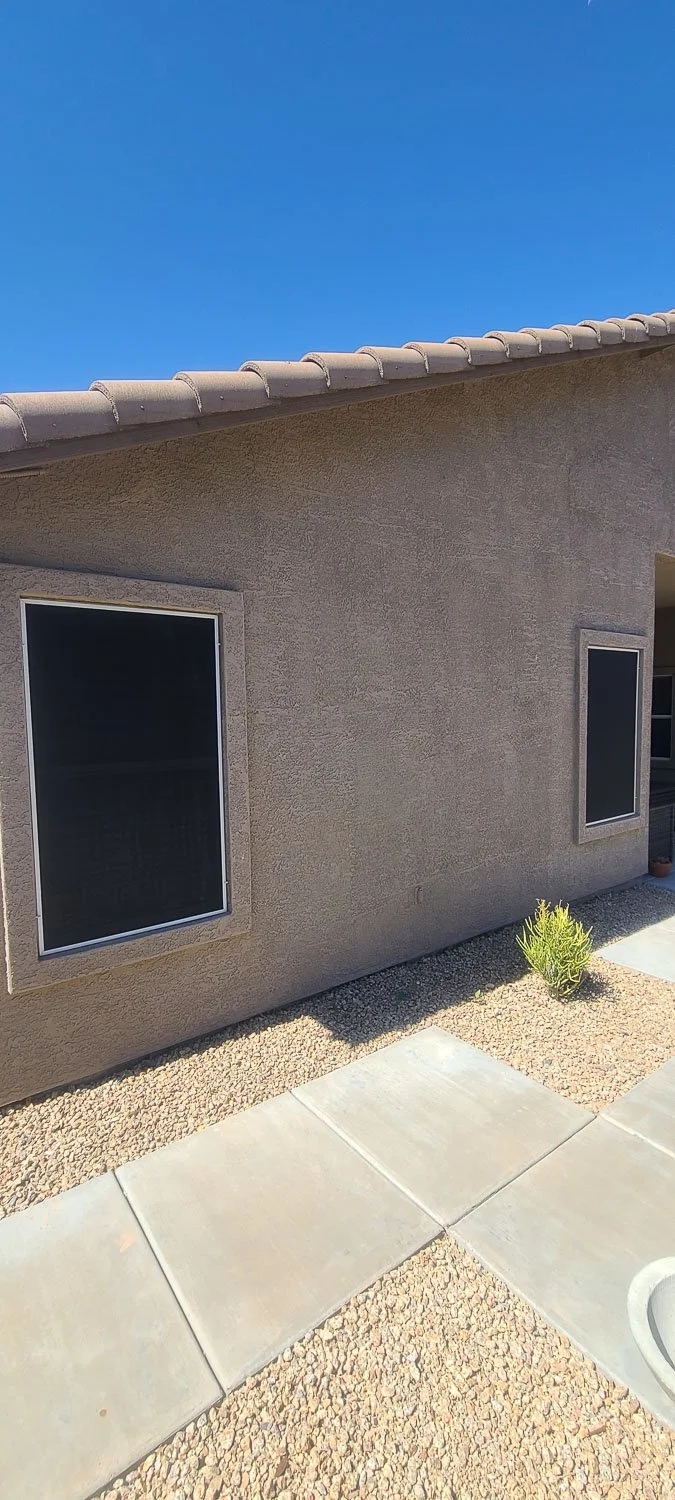 Side view of a house with a textured beige wall, two rectangular windows with dark screens, a sloped roof with beige tiles, a small green bush, concrete sidewalk, and gravel ground beneath.