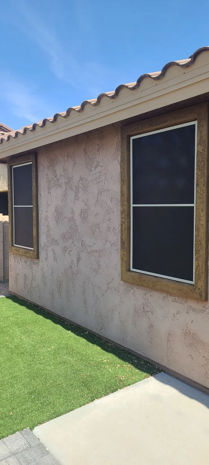 The exterior wall of a house with two closed windows, beige stucco finish, tiled roof, and a small patch of artificial grass in front under a blue sky.
