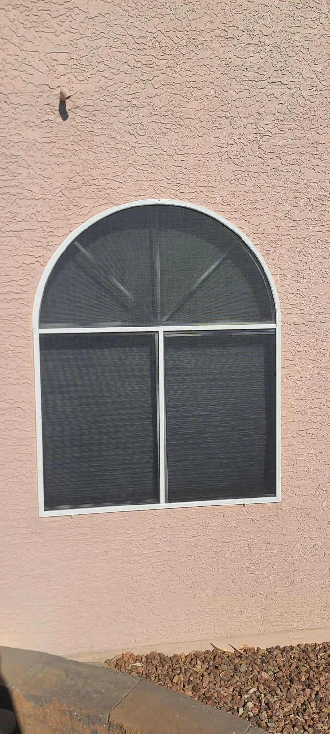 A arched window shade with black mesh screens on a light pink textured wall, with gravel and a curb at the bottom.