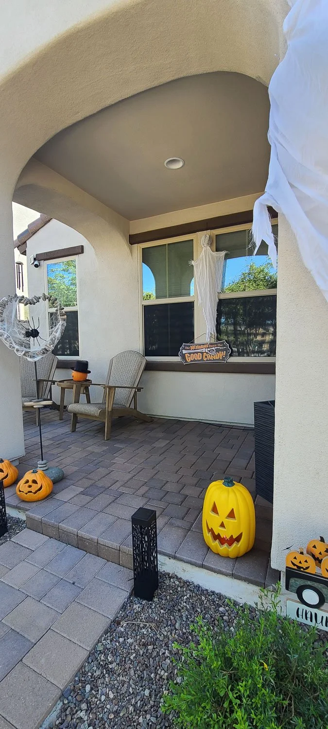 Halloween decorations on a porch, including a large yellow pumpkin with a carved face, smaller pumpkins, a ghost hanging from the ceiling, and a sign that says "The neighbors have the good candy!"