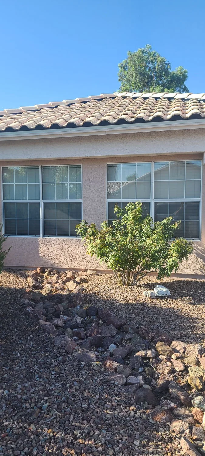 A house with a tiled roof, two large windows with screens, a small bush in front, and a rocky yard under a clear blue sky with a tall tree in the background.