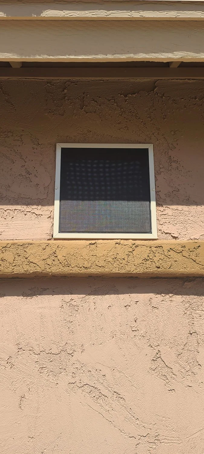 A close-up of a small rectangular window with a screen on a textured pinkish-beige stucco wall, with part of a white window frame visible.