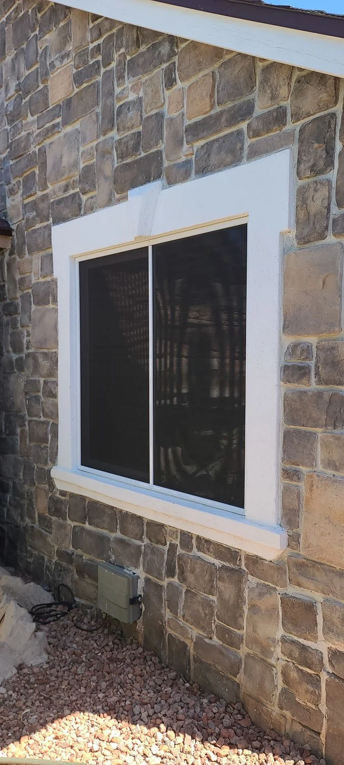 Exterior view of a stone house wall with a window and a screened window door, surrounded by gravel and construction materials.