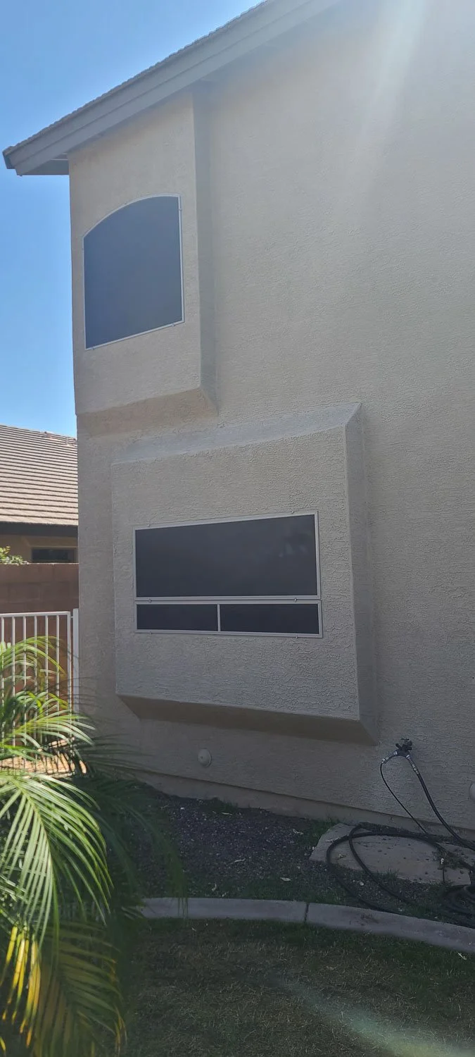 Side view of a two-story house with beige stucco exterior, two large window screens, some greenery, and a garden hose coiled on the ground.