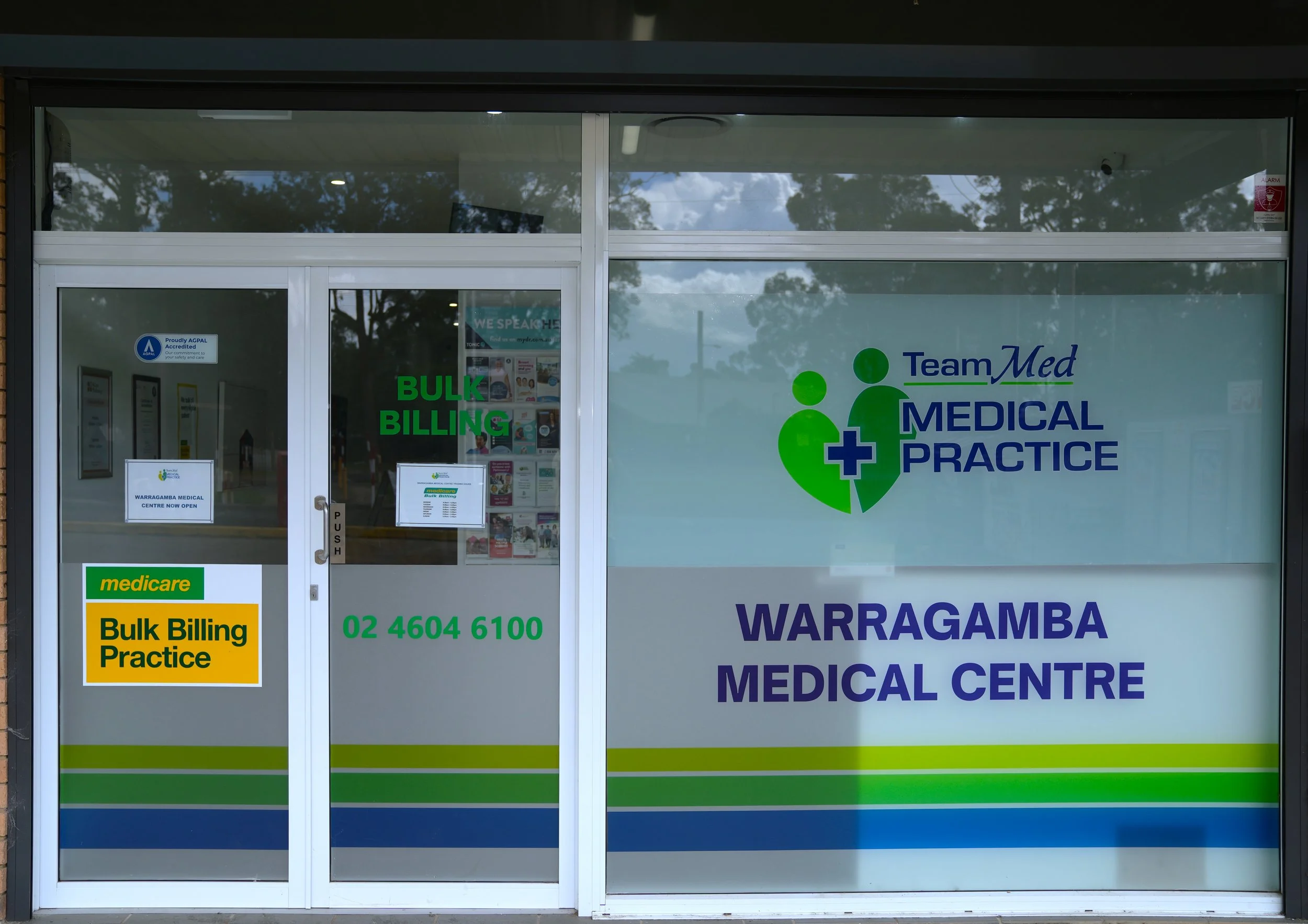 The storefront of a medical practice with clear glass doors and windows, displaying signs for bulk billing, Medicare, and the Warragamba Medical Centre. Reflective trees and sky can be seen through the glass.