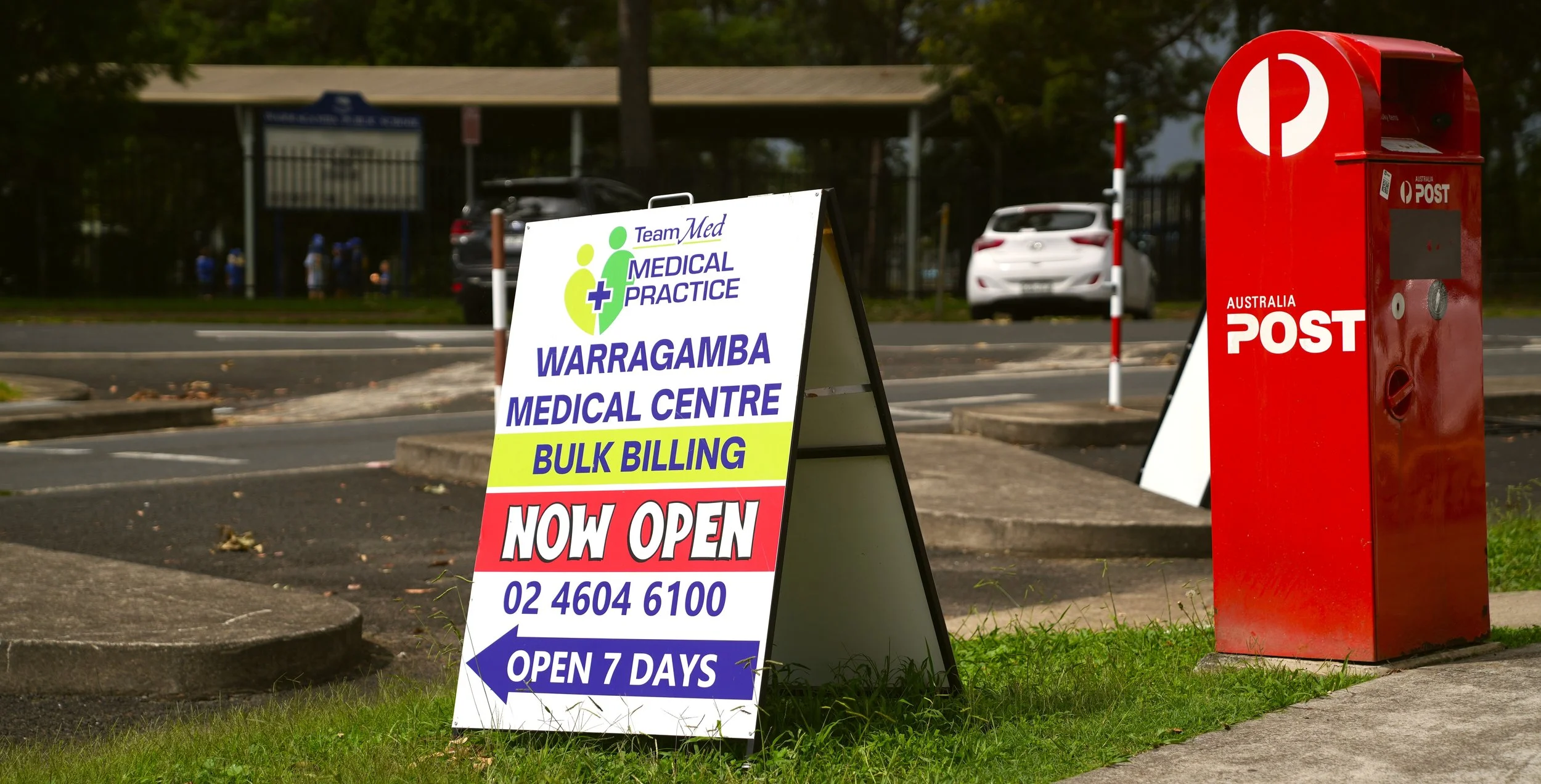 A red Australia Post mailbox and a white signboard for Warragamba Medical Centre, indicating bulk billing and 24/7 opening, with a parking lot and trees in the background.