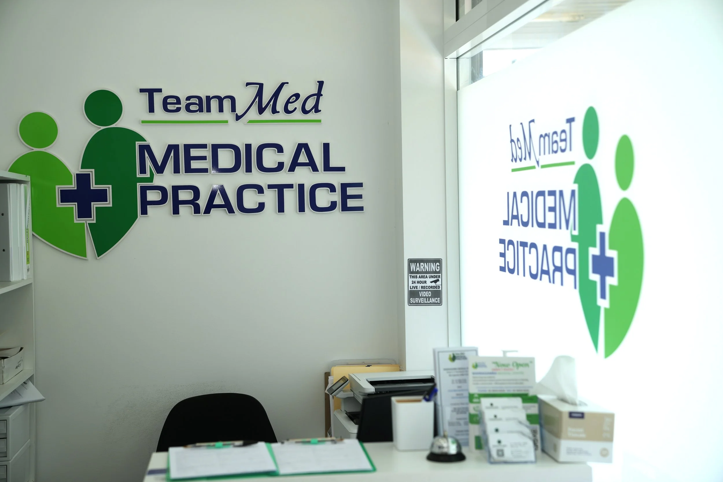 Reception area of a medical practice with the name 'Team Med Medical Practice' displayed on the wall, featuring a logo with green circles and a caret symbol, near a window with sunlight streaming in. There are office supplies and promotional materials on the desk.