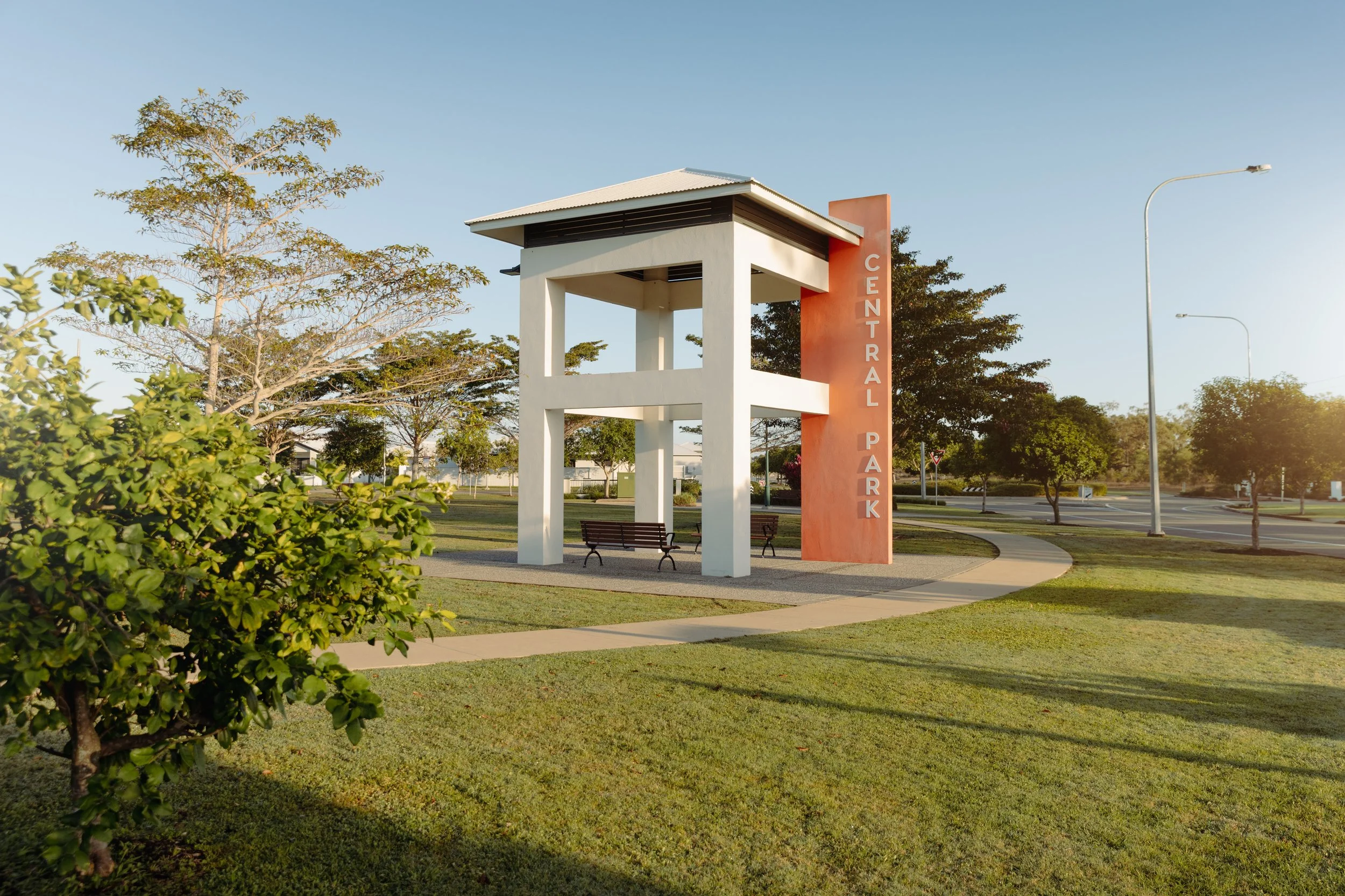 Photo of a park entrance structure with a bench underneath. The structure has white pillars and a red vertical sign with white letters that read 'CENTRAL PARK'. The park has green grass, trees, a paved walkway, and streetlights, with a clear blue sky