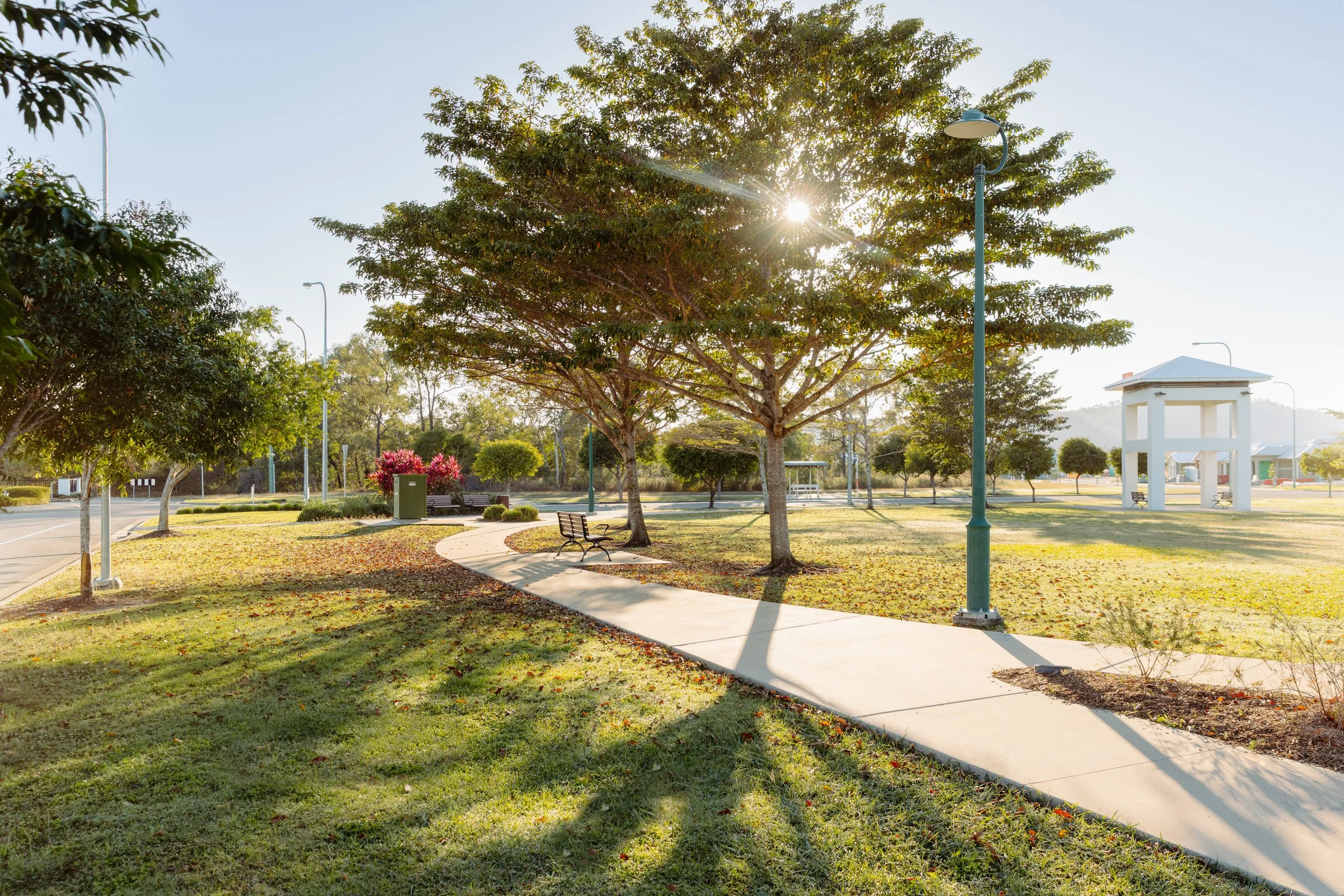 A sunny park scene with trees, a winding walkway, benches, and a white gazebo.