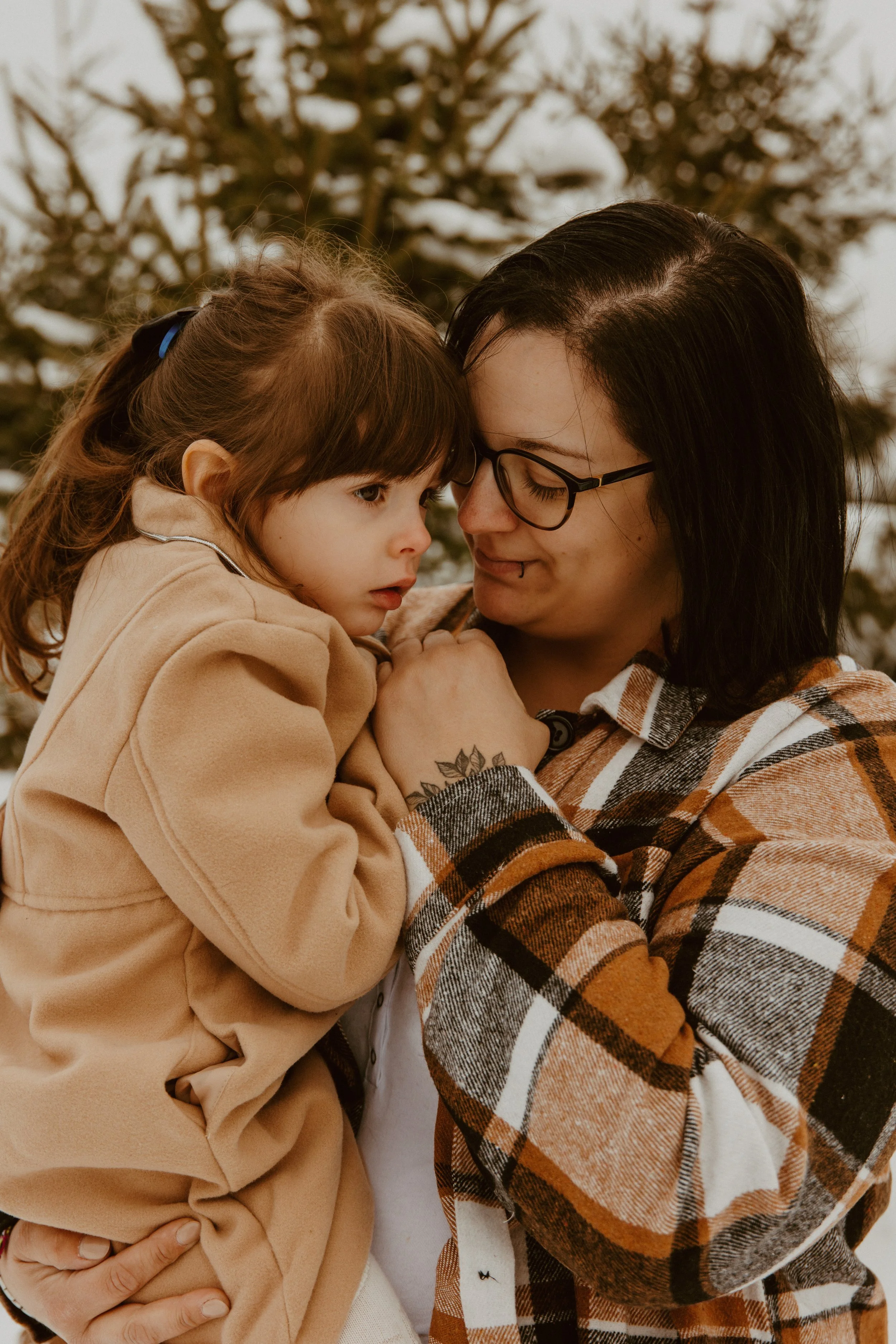 Une femme avec des lunettes et des cheveux noirs tenant une jeune fille aux cheveux bruns, qui porte un manteau beige, dans ses bras. Ils se regardent tendrement dans un environnement extérieur avec des arbres en arrière-plan.