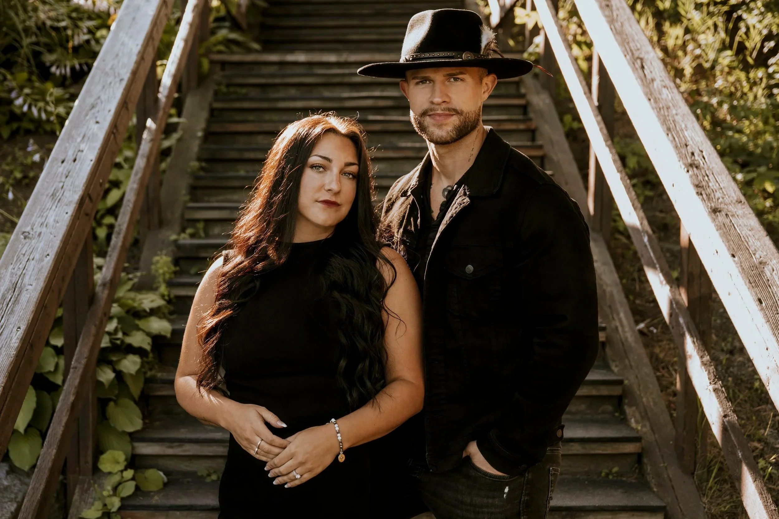 Un homme et une femme posent côte à côte sur un escalier en bois, entourés de végétation, lors d'une séance photo en extérieur, tous deux habillés en noir.