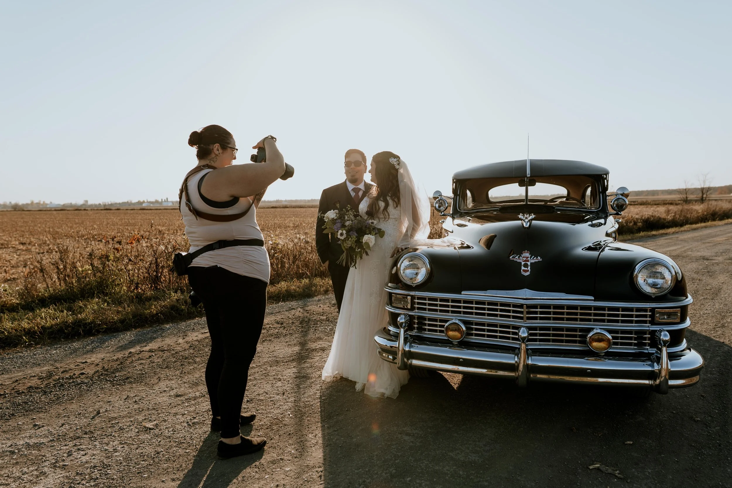 Un couple de mariés en costume dans une campagne ensoleillée, posant pour une photo avec une voiture ancienne noire, une femme avec une robe de mariée et un homme en costume, une femme prenant la photo avec un appareil photo.