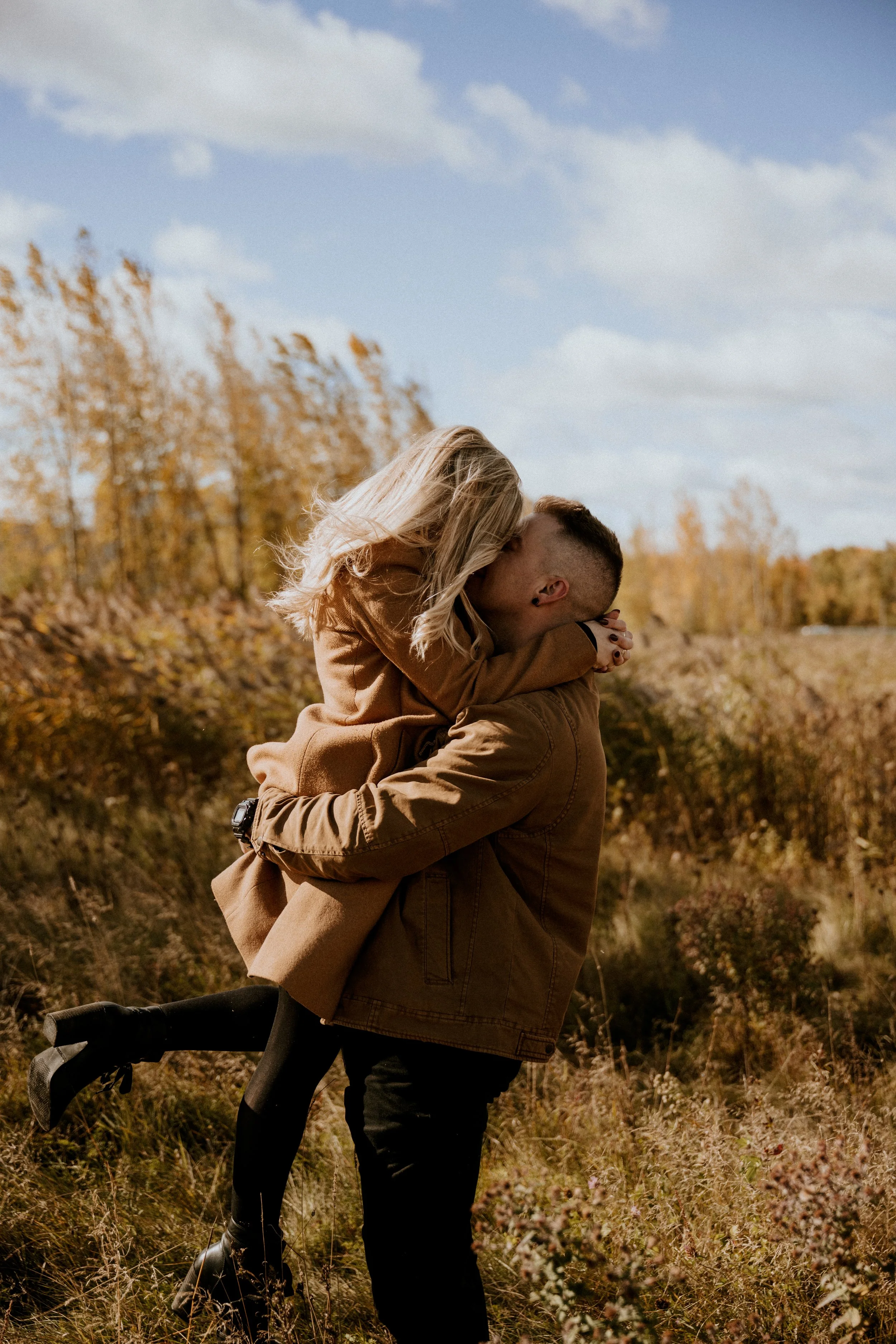 Un couple fusionnel, l'homme portant la femme sur ses épaules dans un paysage automnal avec des arbres aux couleurs d'automne et un ciel partiellement nuageux.