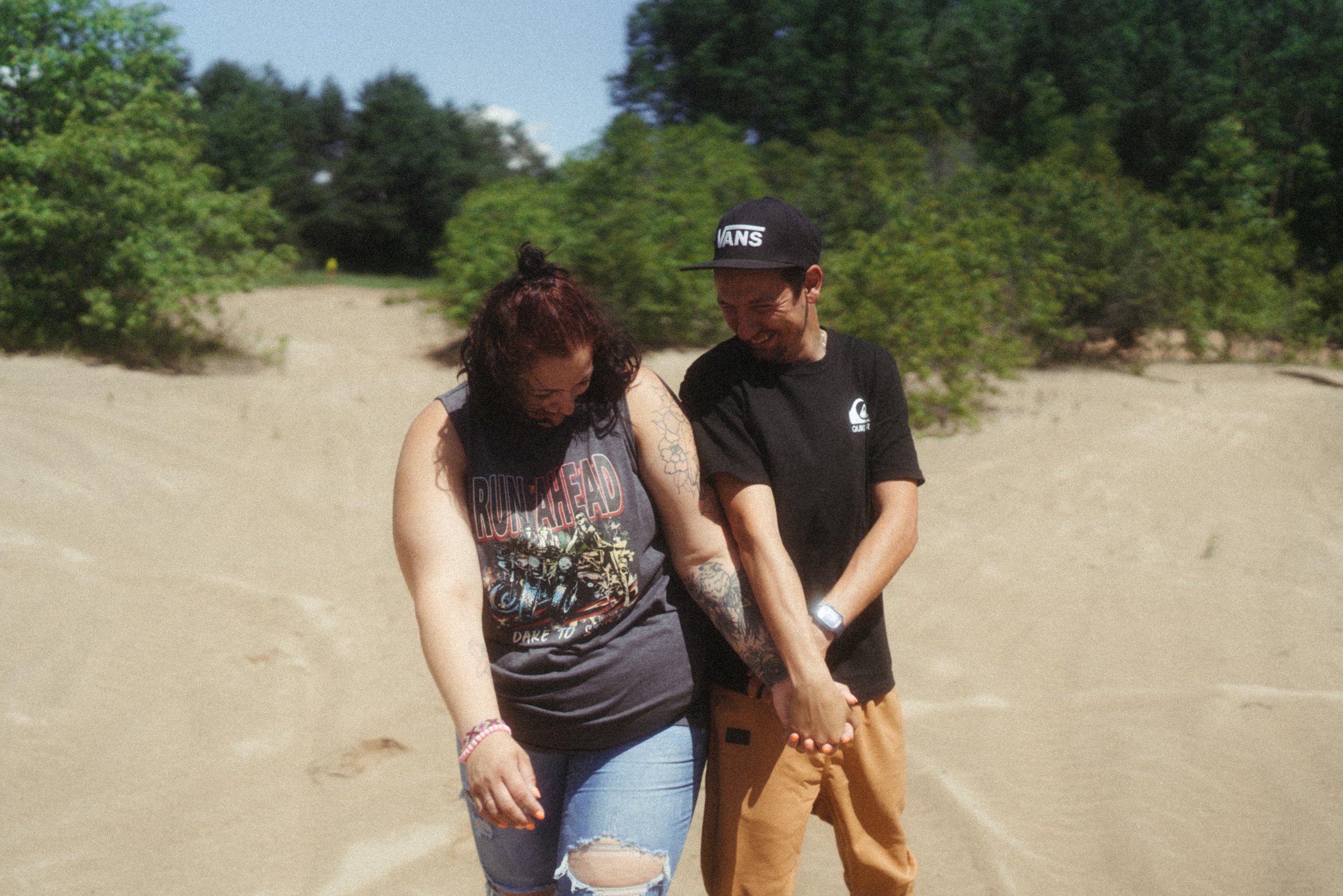 Deux personnes marchent main dans la main sur une plage en sable, entourées de végétation verte et de buissons, par une journée ensoleillée.