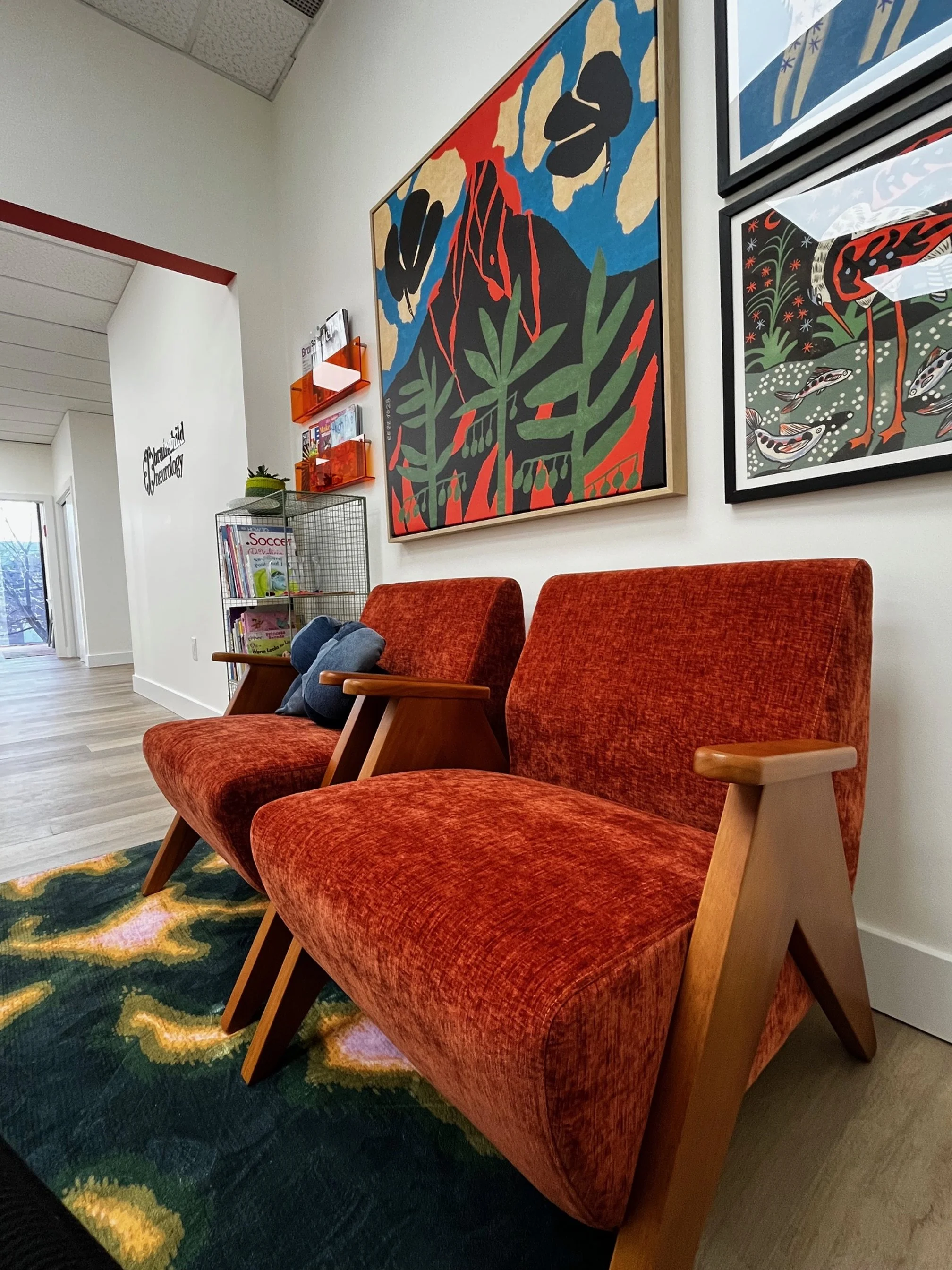 Waiting area with two red-orange upholstered chairs with wooden armrests, colorful artworks on the white wall behind, and a colorful rug on the wooden floor.