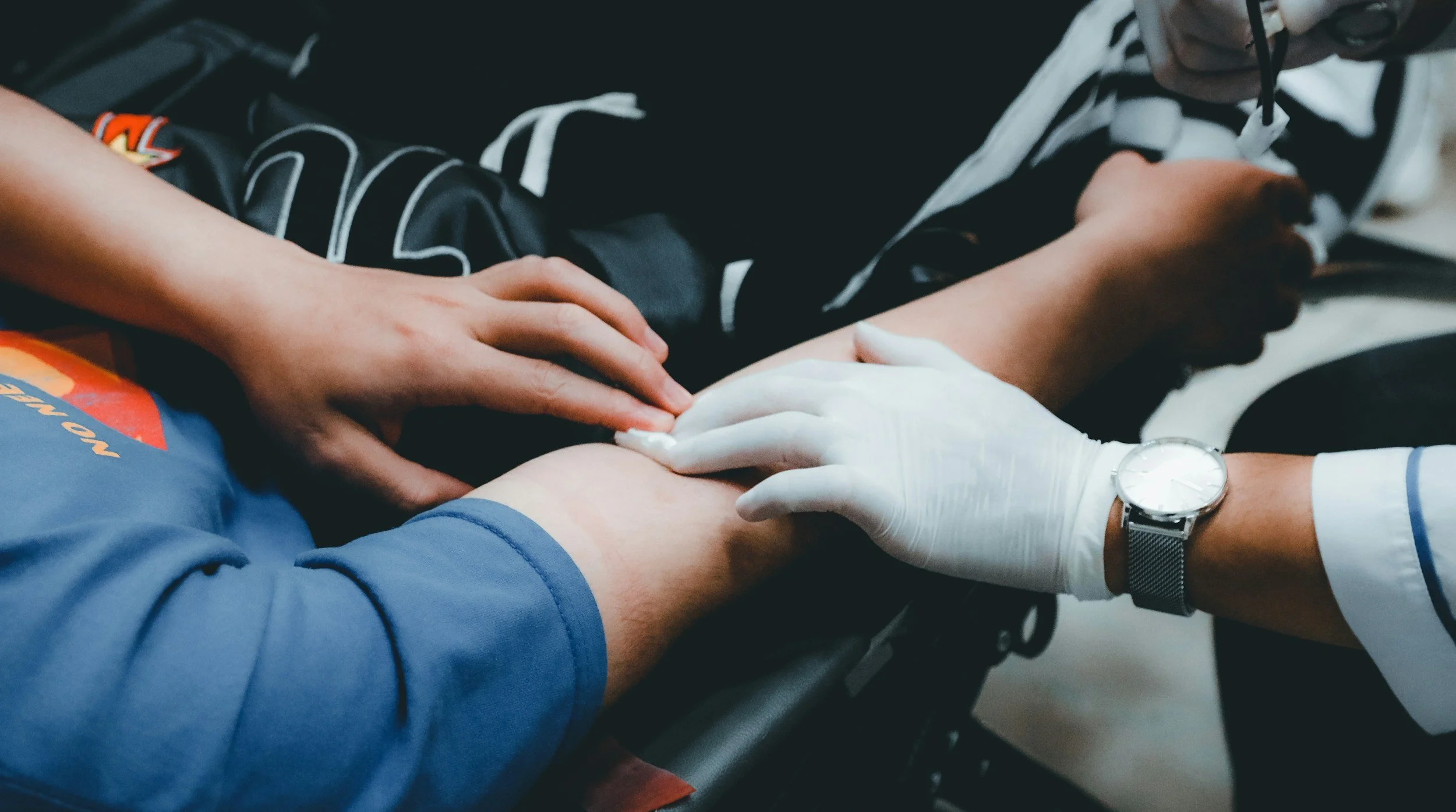 A person in a blue shirt receives a medical injection in their arm from healthcare worker wearing gloves and a watch.