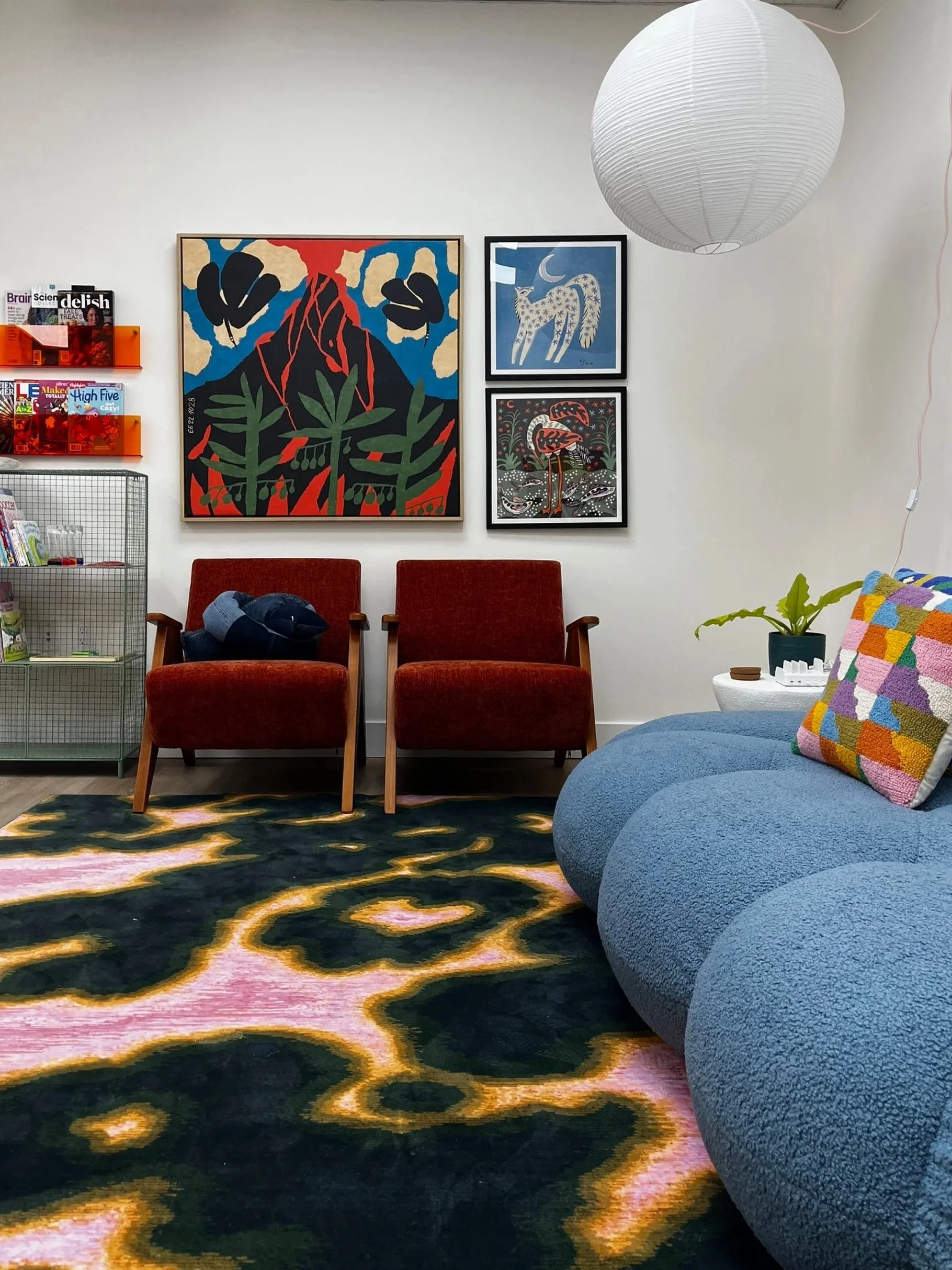 A cozy waiting room with red chairs, a colorful patterned carpet, a blue sofa with a multicolored pillow, a white side table with a plant, framed artwork on the wall, and a large white paper lantern hanging from the ceiling.