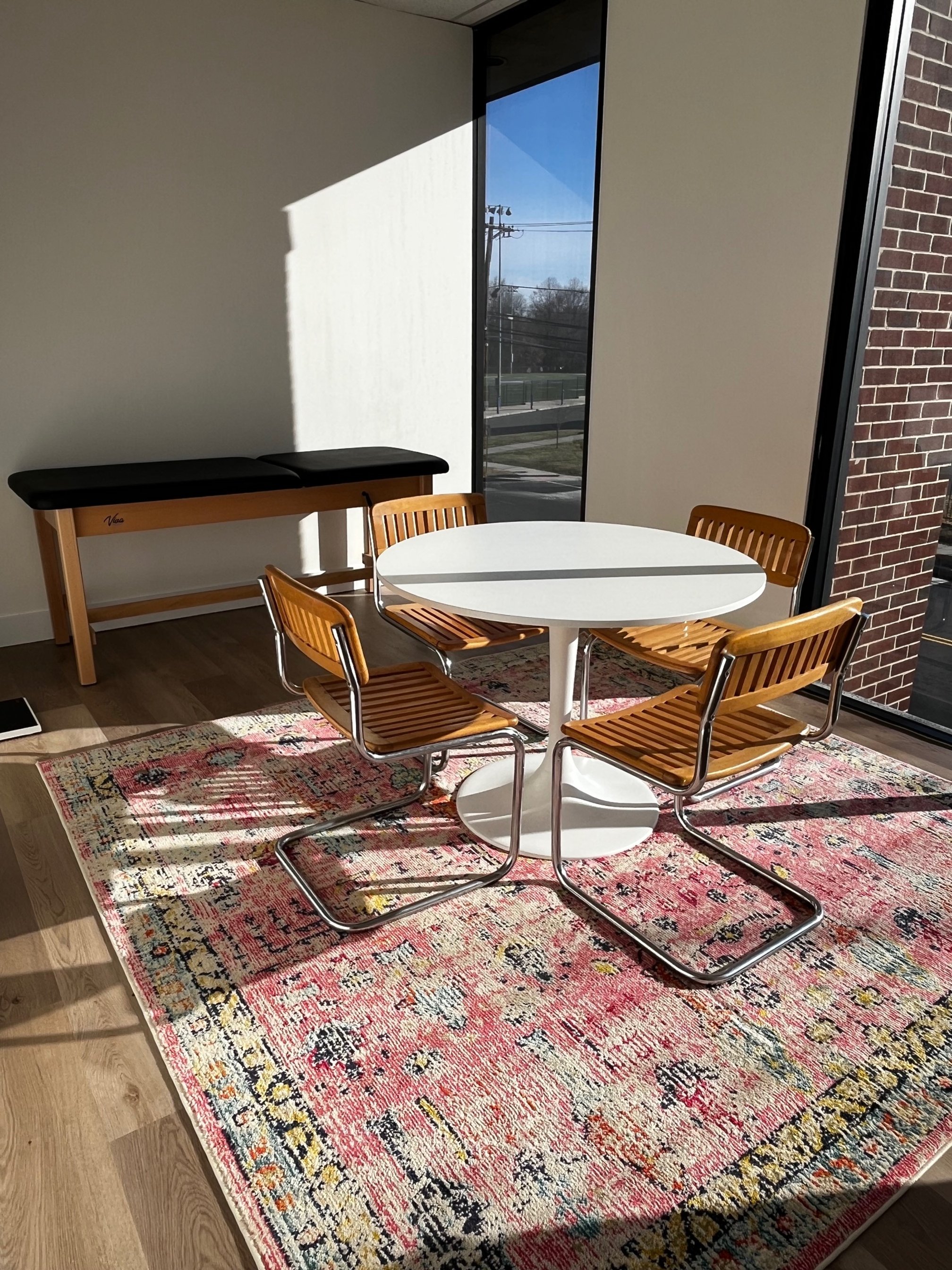 A small round white table in our exam room with four wooden chairs on a colorful patterned rug. Sunlight is streaming in through large glass windows, casting shadows inside a modern room with hardwood flooring and a brick wall.