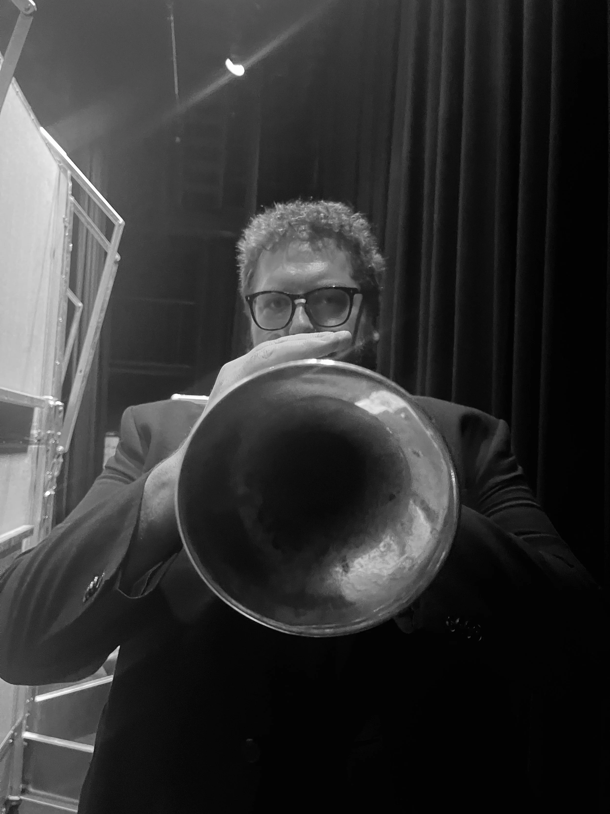 A man with glasses and curly hair playing a trumpet, seen in a mirror with stage curtains behind him.