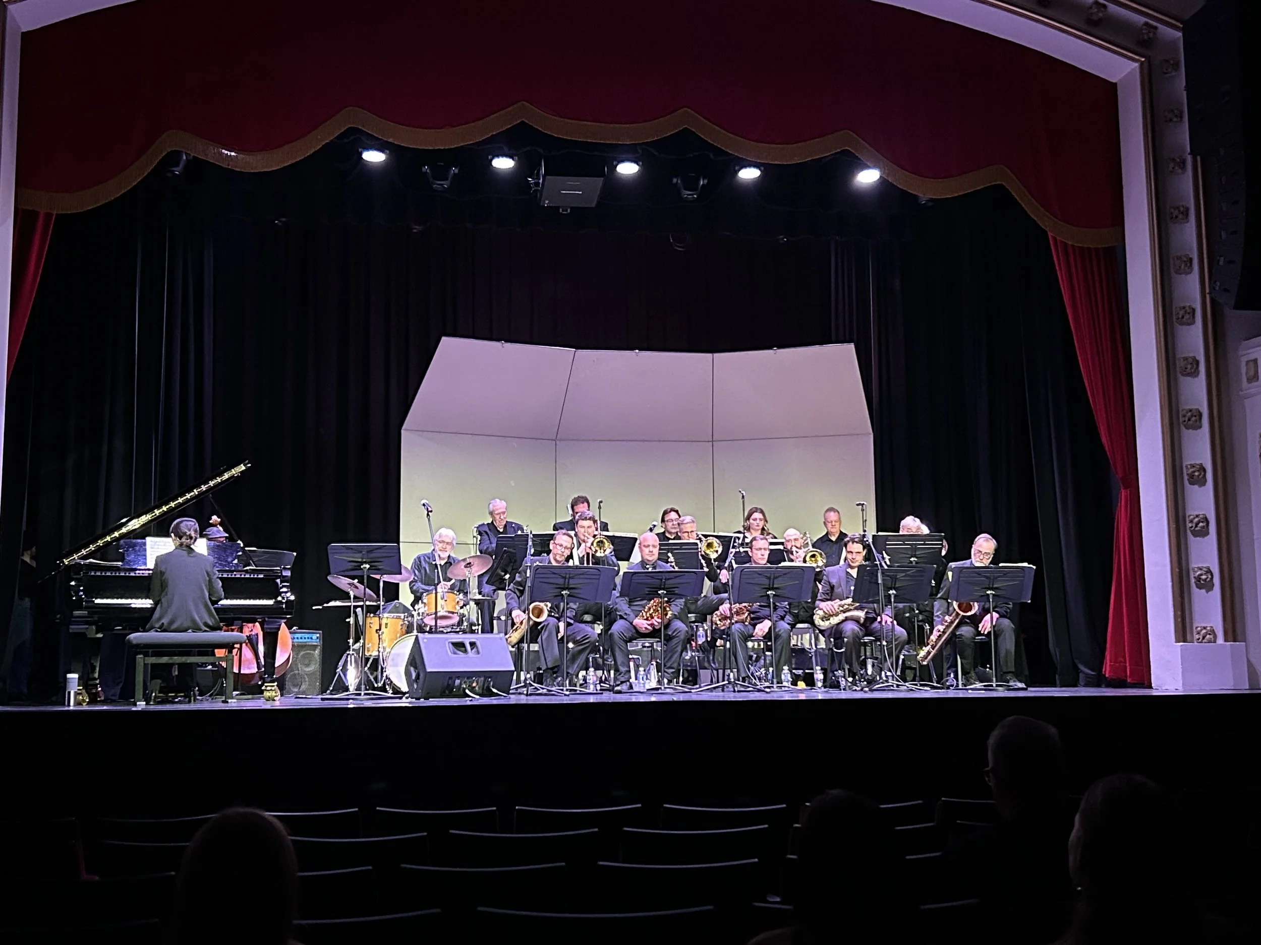 Jazz band performs on stage in a theater with red curtains and black backdrop, featuring a pianist, drummer, and multiple wind instrument players.