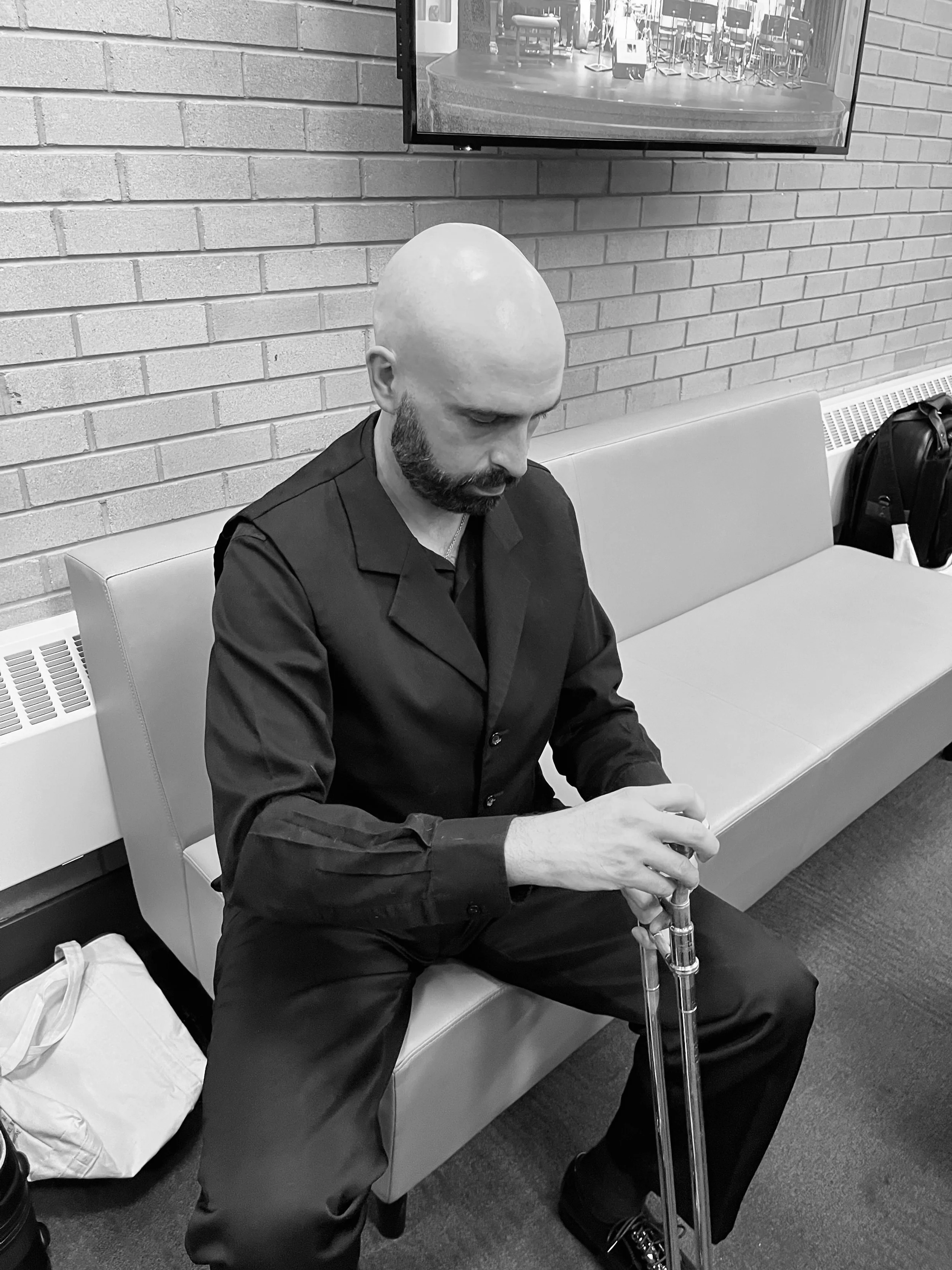 A bald man with a beard sitting on a bench in a waiting area, holding a cane, with a brick wall and television in the background.