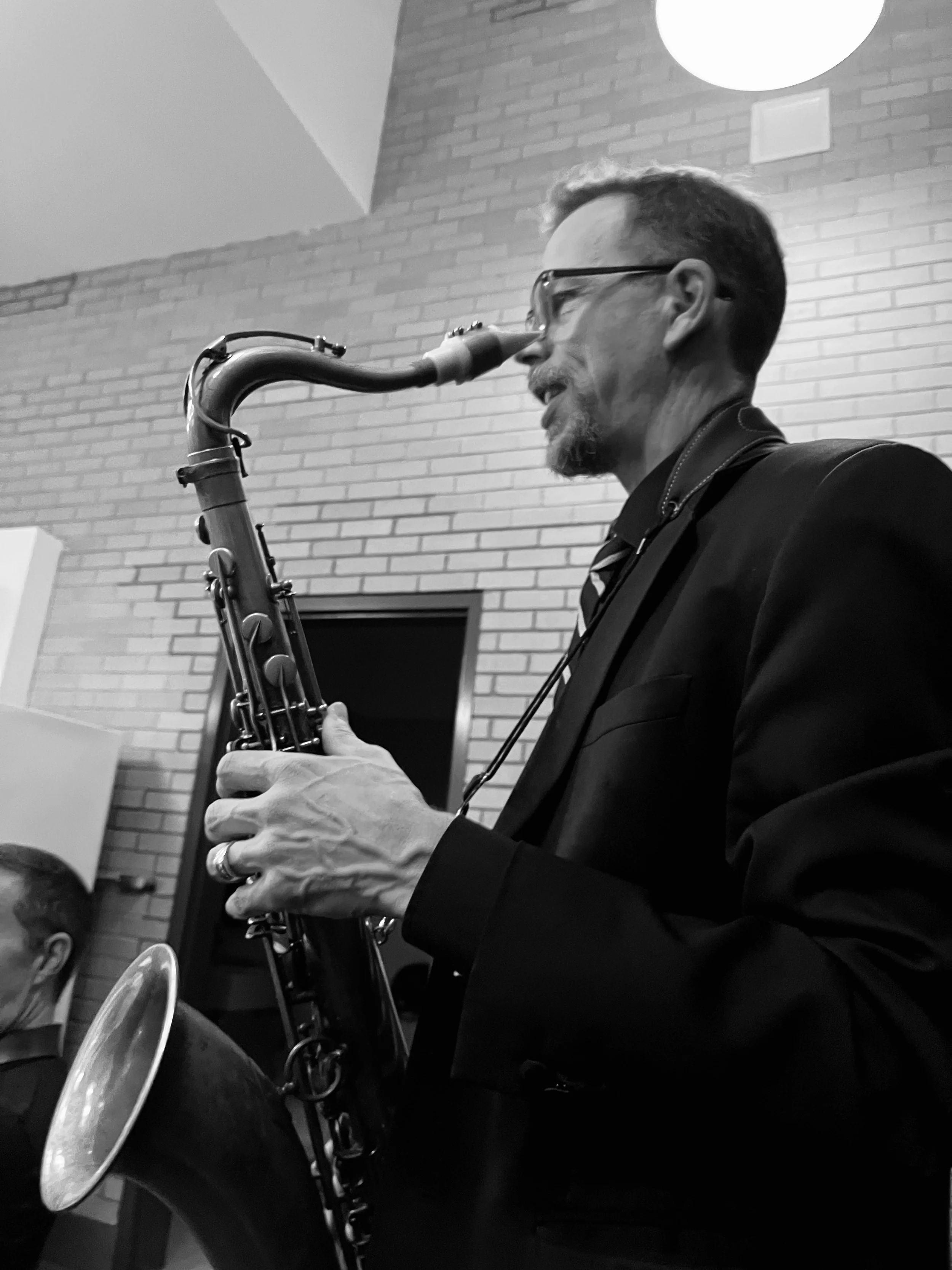 A man in a suit playing a saxophone indoors, with brick walls in the background.