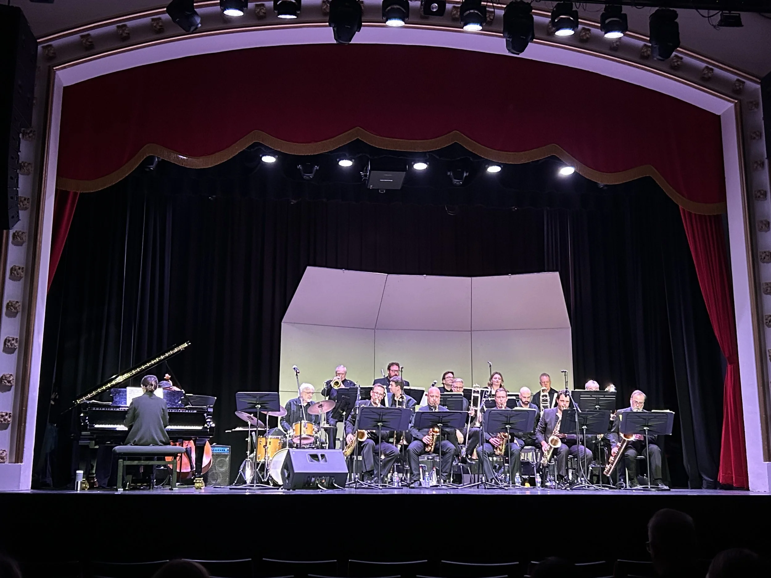 Jazz band performing on stage with various instruments, including piano, drums, and brass, in a theater with red curtains.