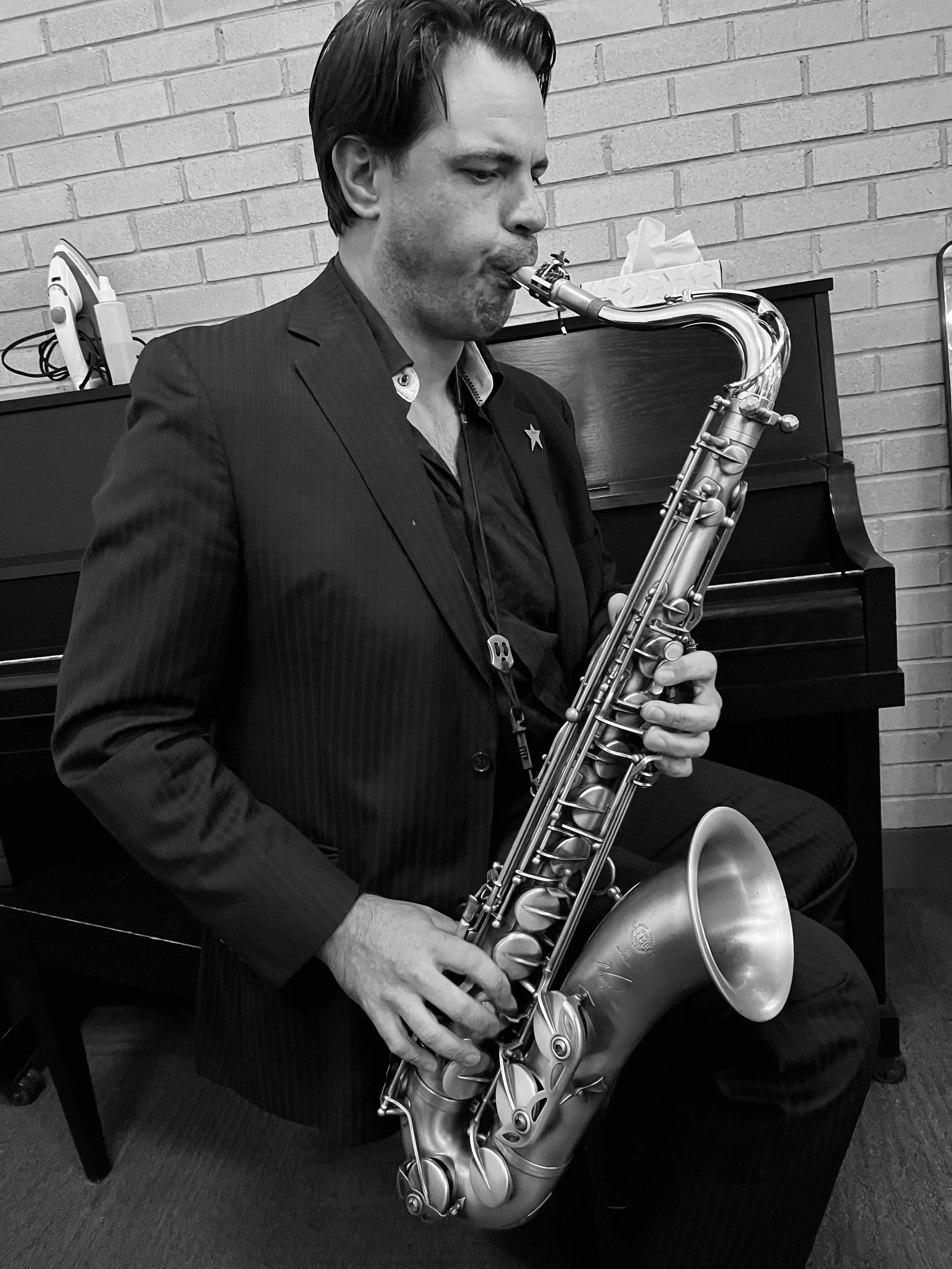 A man in a suit playing a saxophone indoors, sitting in front of a piano against a brick wall.
