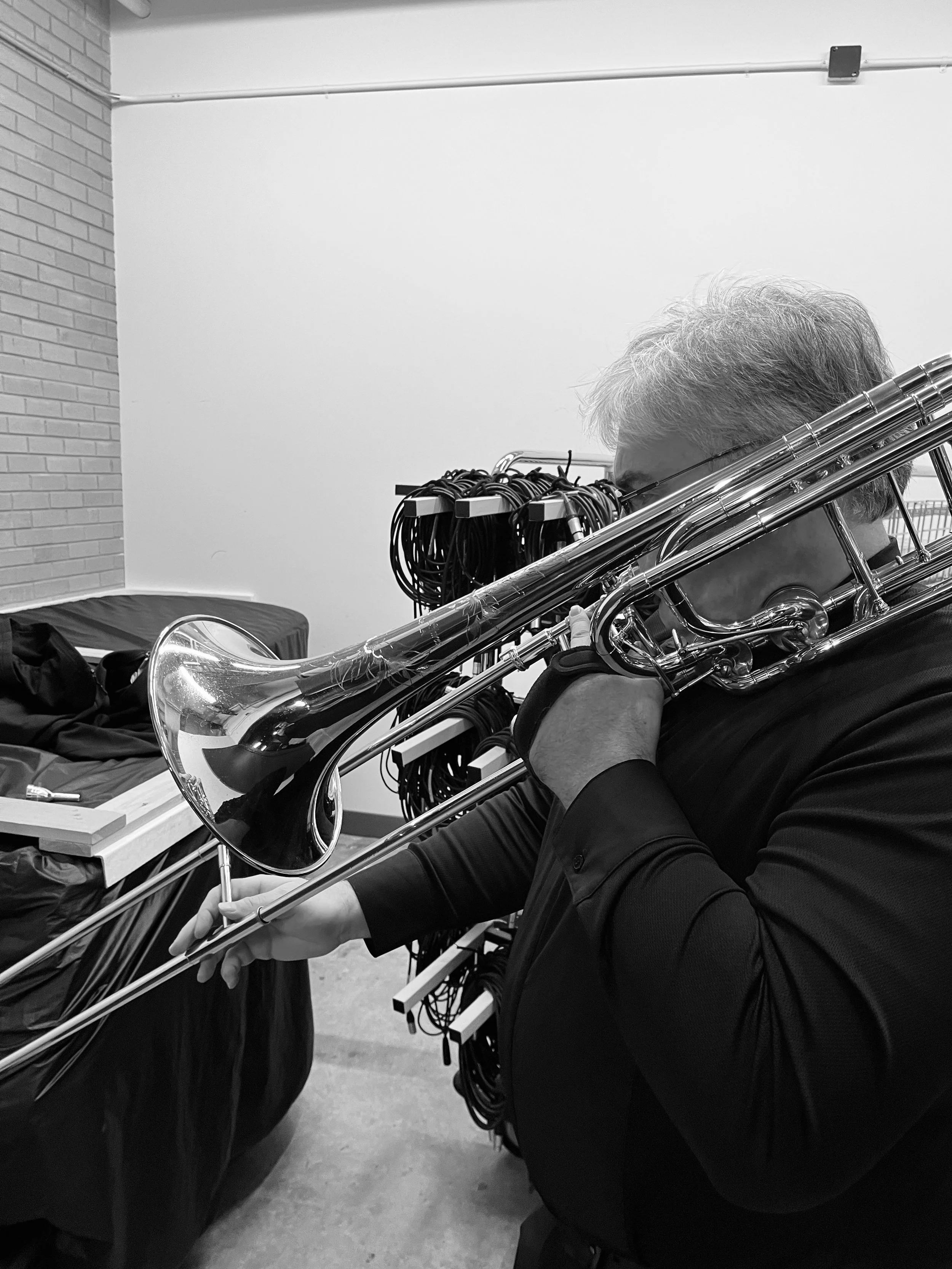 A person with gray hair holds a trumpet resting on their shoulder in a room, with audio cables and equipment in the background.