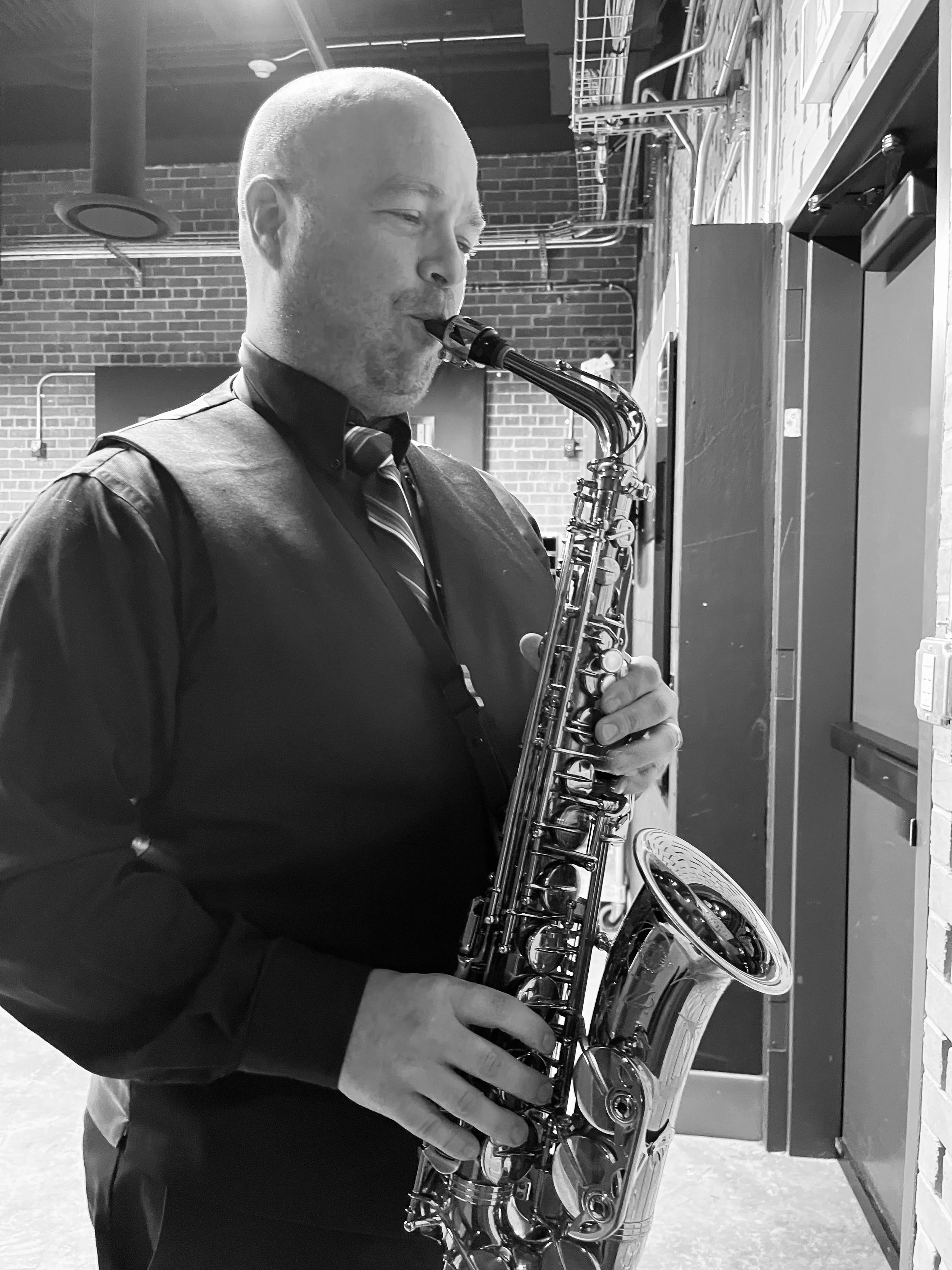 A man in formal attire, wearing a vest, shirt, and tie, playing a saxophone in an indoor setting with brick walls and exposed pipes.