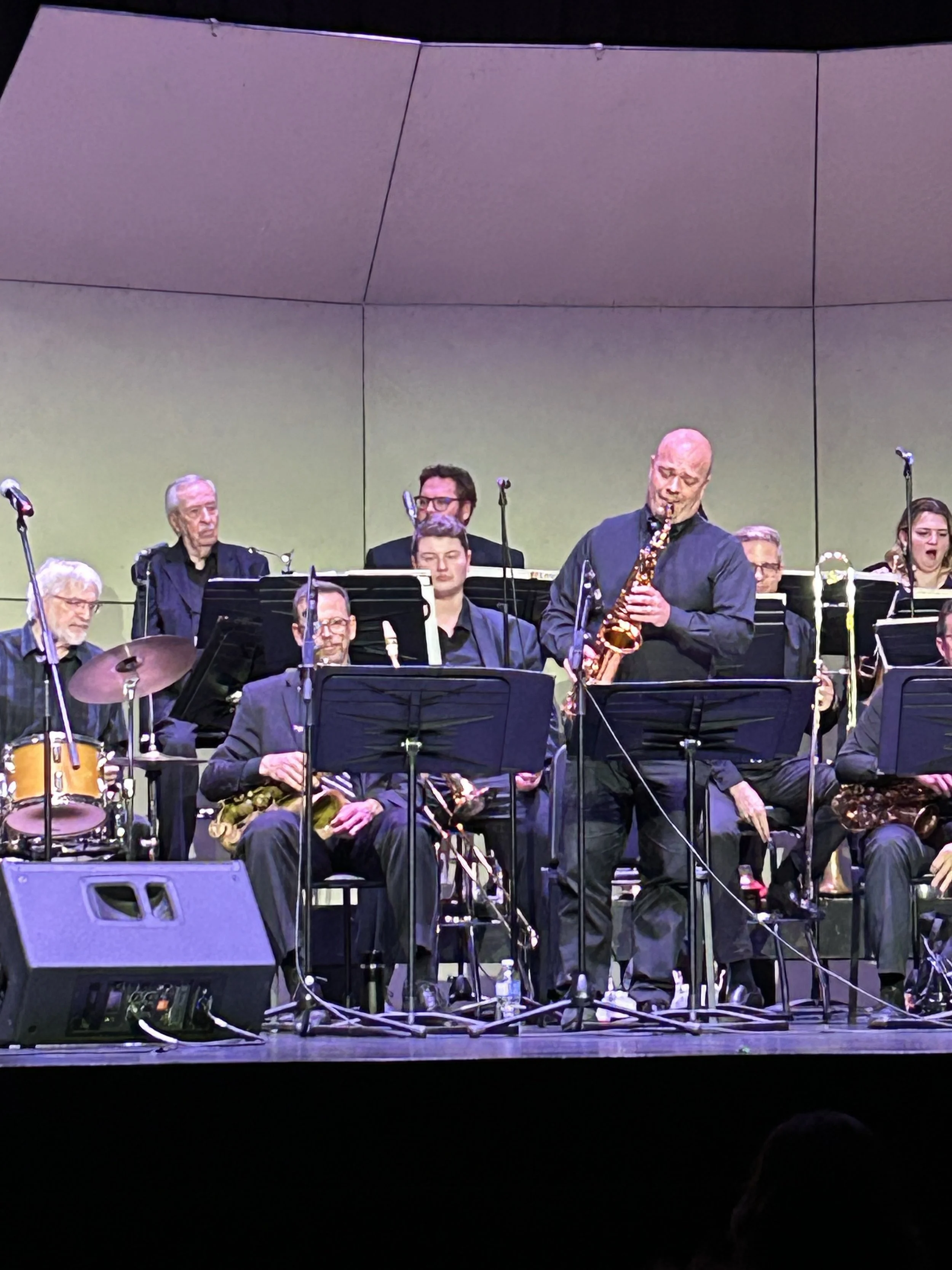 Jazz band performing on stage with musicians playing drums, saxophones, and other instruments.