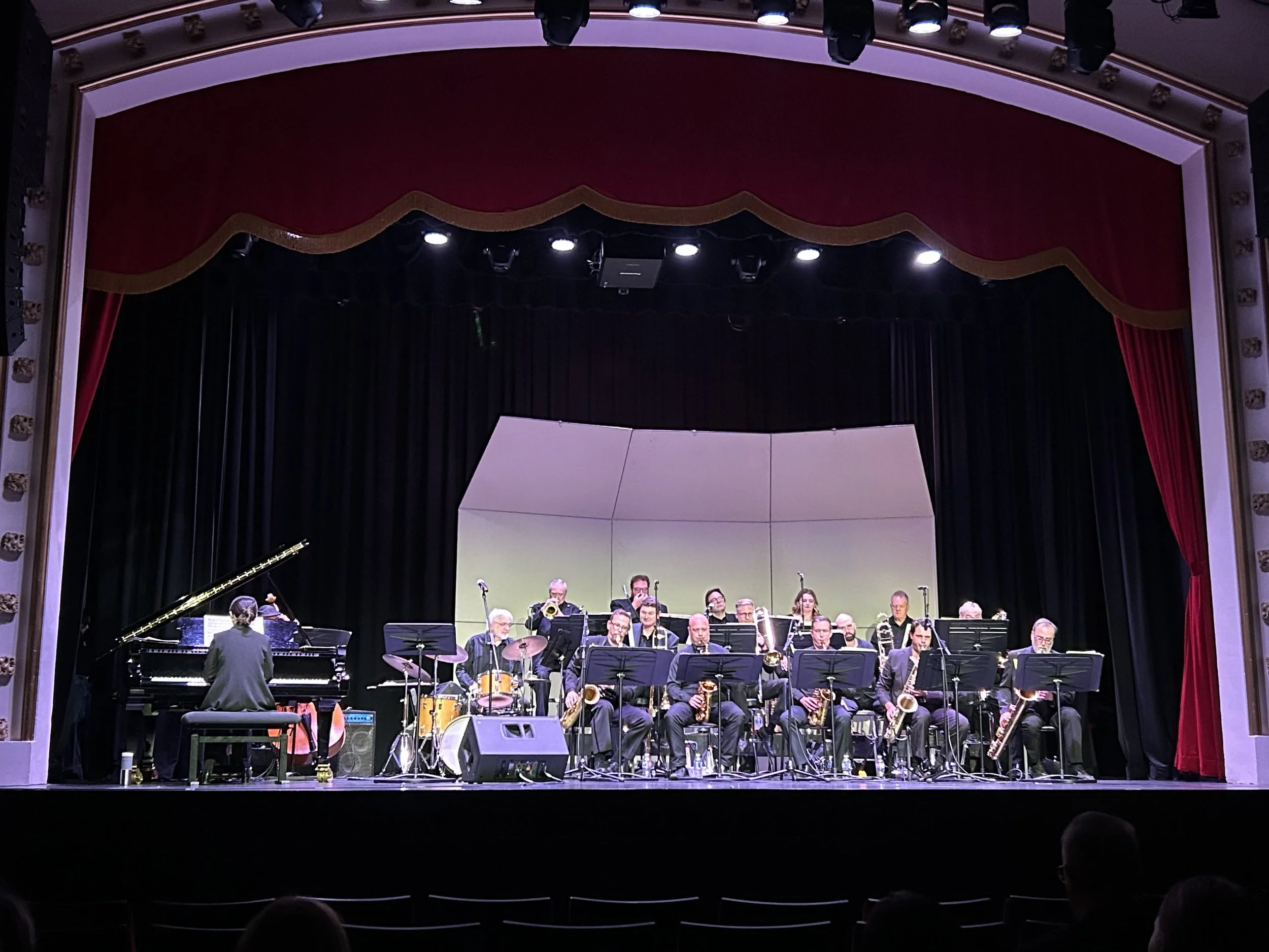 Jazz band performing on stage with a pianist and various wind instruments, in a theater with a red curtain and black backdrop.