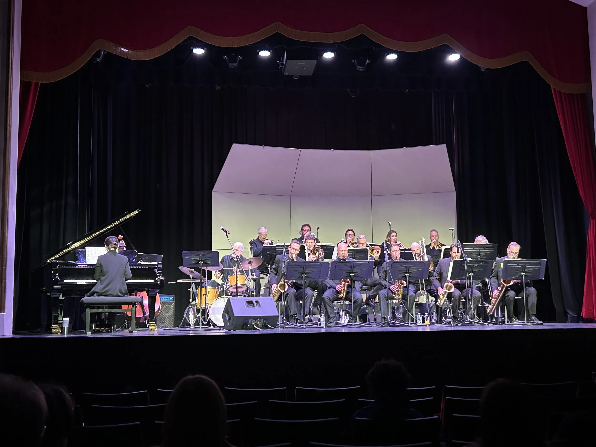Big band jazz orchestra performing on stage with a pianist, trumpet players, saxophonists, and percussion, in a theater with dark curtains and stage lighting.