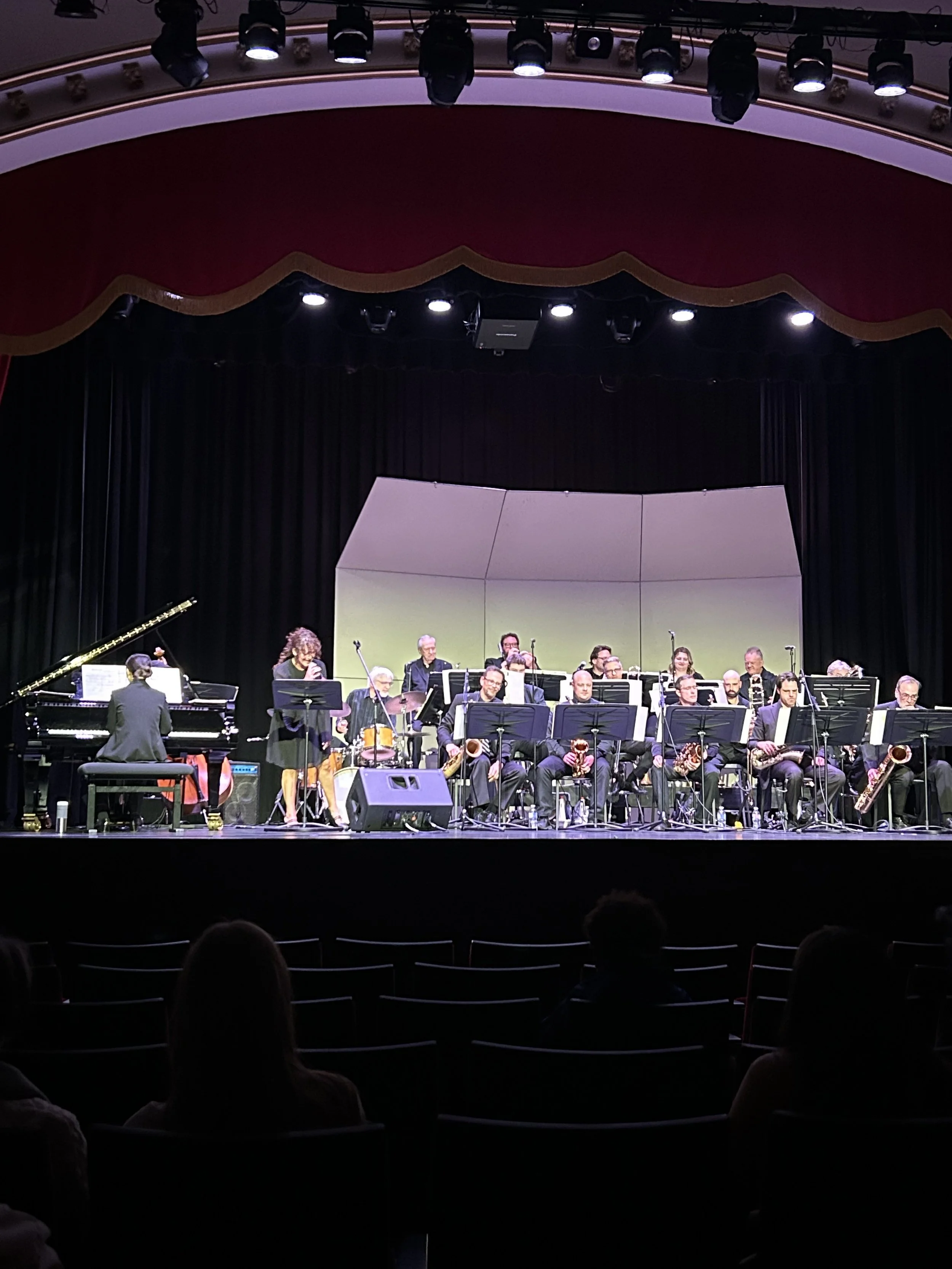 Large jazz band on stage with a pianist, singer, and musicians playing various instruments including saxophones and drums, illuminated under stage lights in a theater with a red curtain and audience in front.