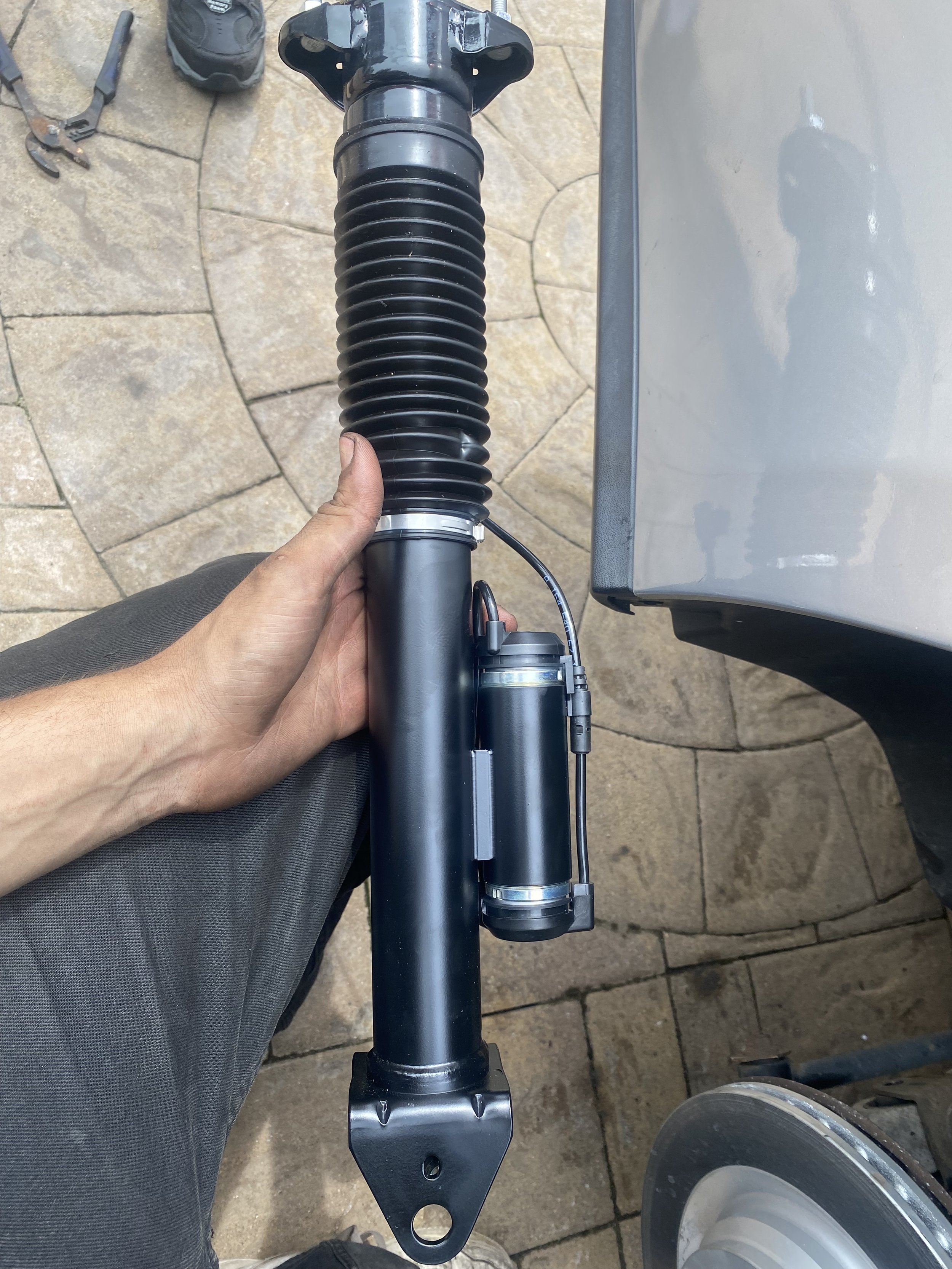 Close-up of a person's hand holding a motorcycle front fork suspension, with a brake disc and concrete pavement in the background.