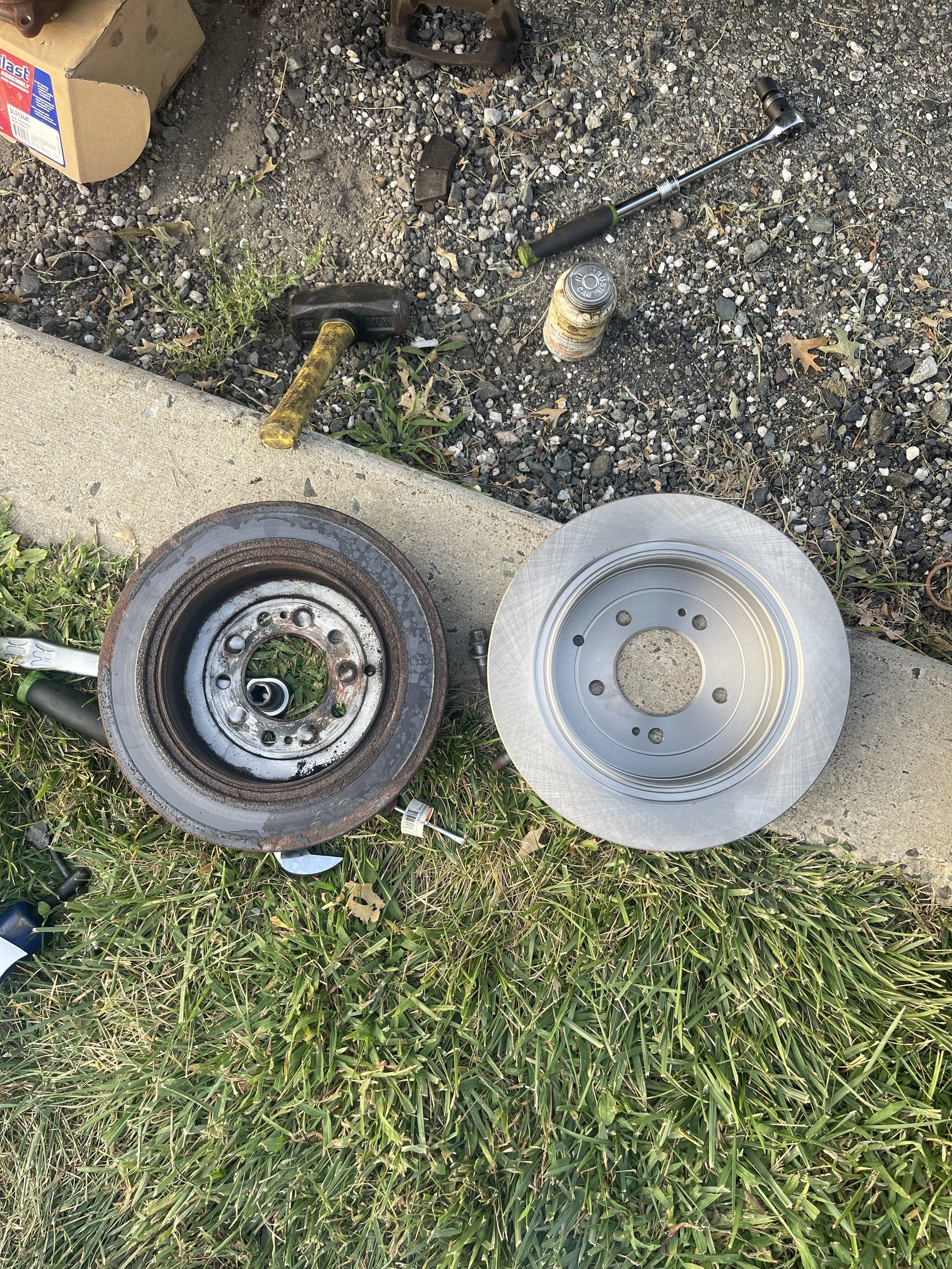Disassembled automotive brake rotor with one rusty and one clean rotor placed on grass and concrete, with tools and parts nearby.