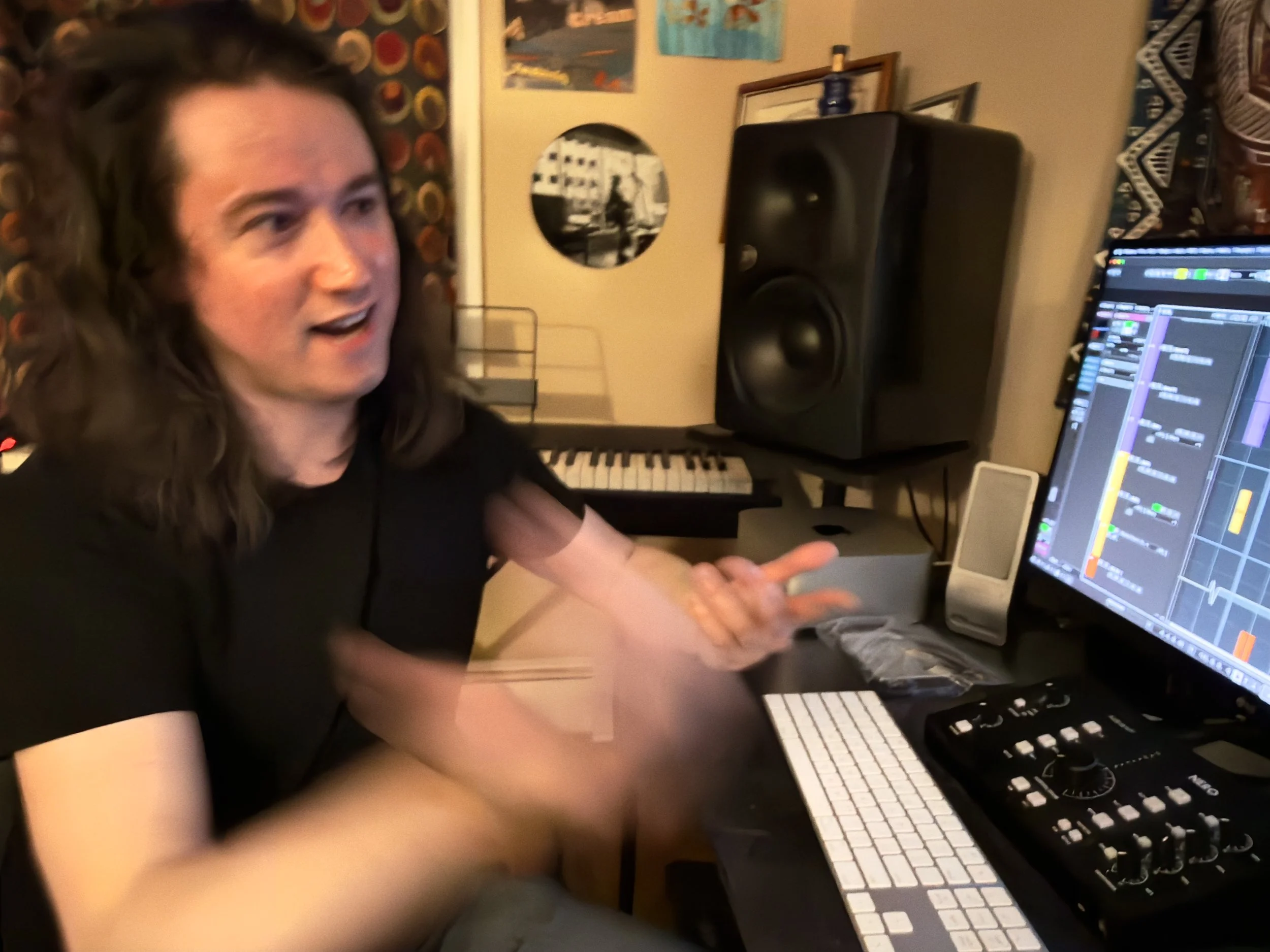 A person with long curly hair, wearing a black shirt, is sitting at a desk in front of a computer. The computer screen shows digital audio workstation software. Behind the person, there are large speakers, a keyboard, and various pieces of wall art in the room.
