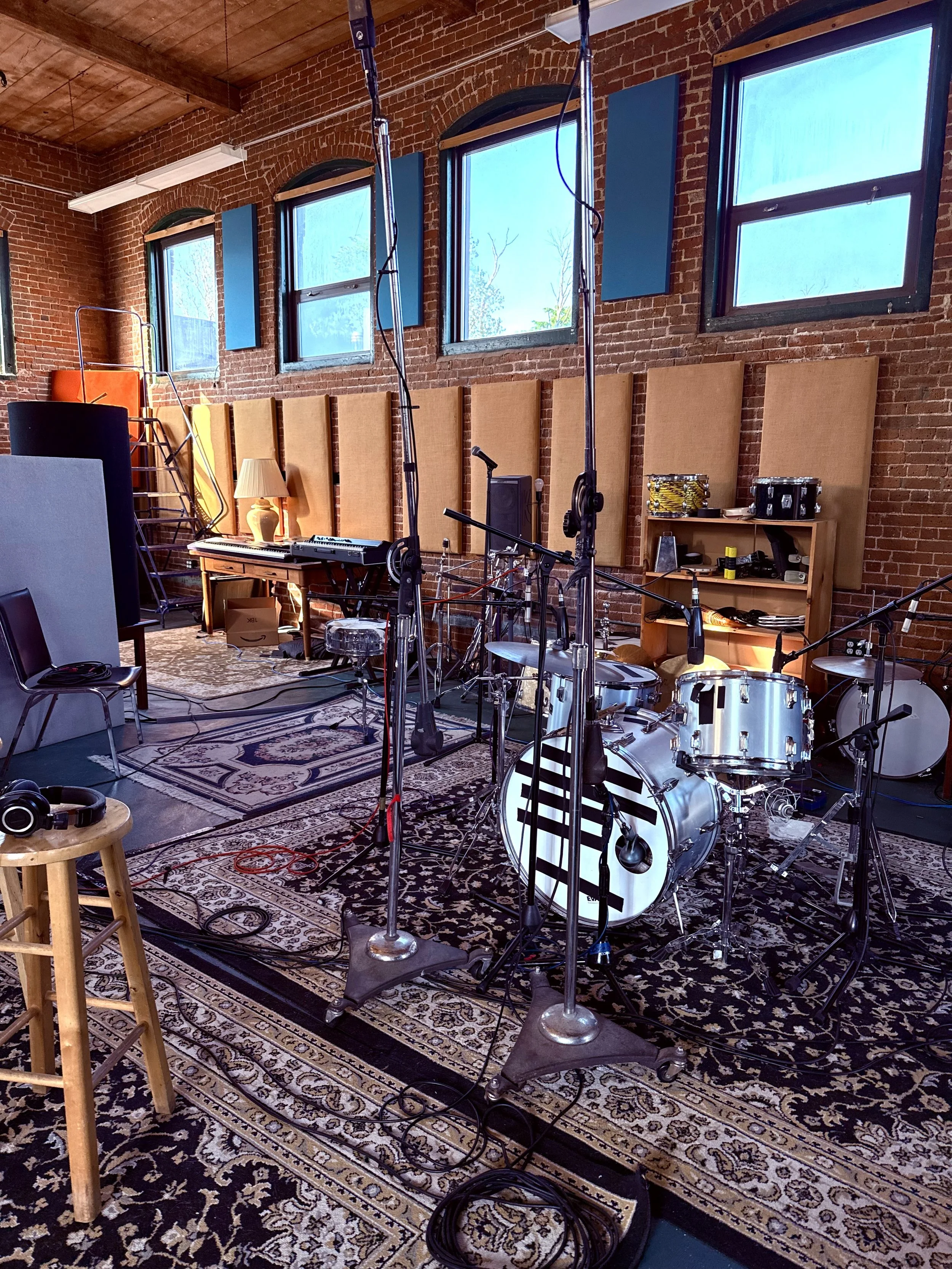 Music studio with multiple musical instruments including drums, keyboard, and microphones, set up in front of a brick wall with windows, acoustic panels, and sound equipment.