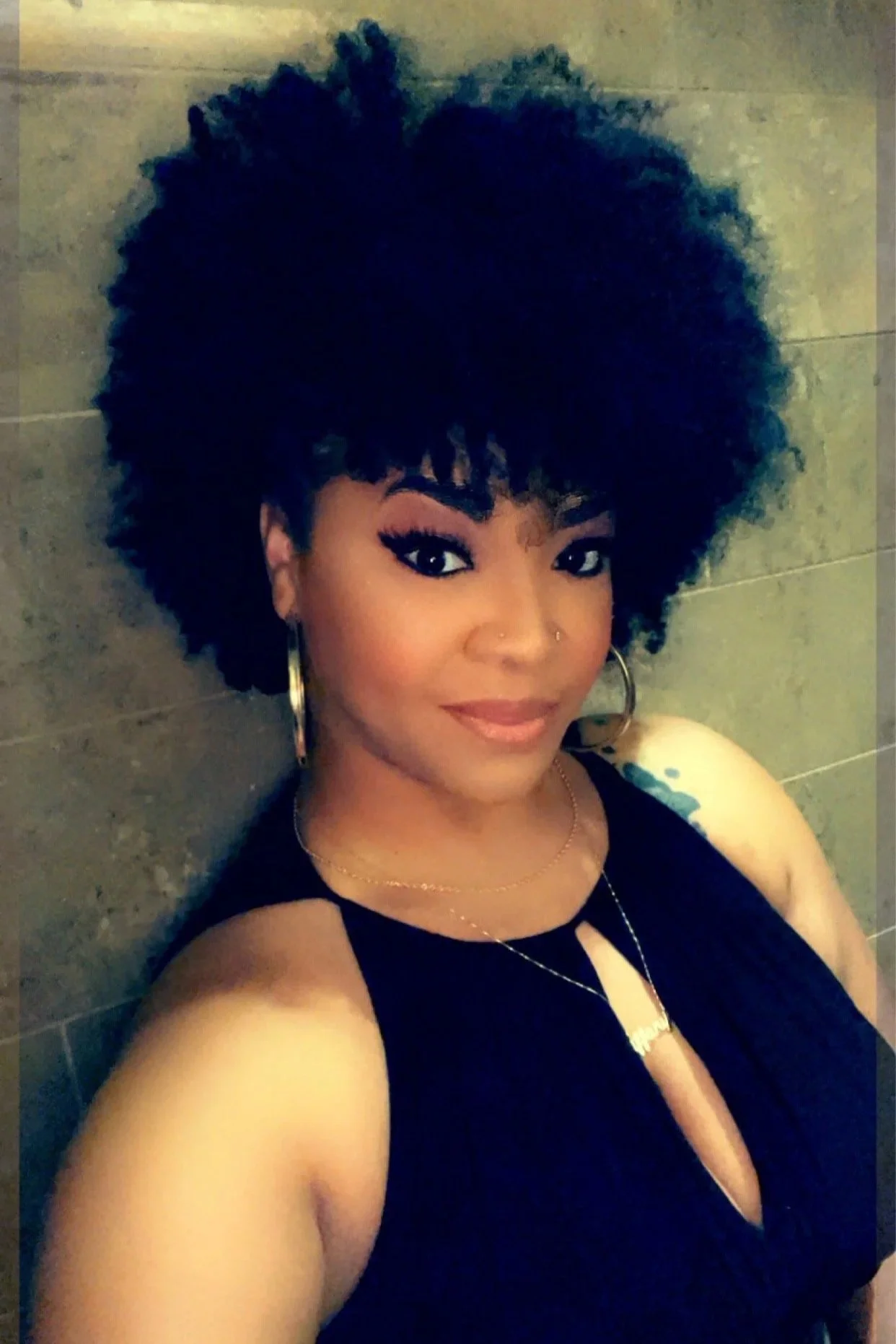 A woman with dark skin and curly afro hairstyle taking a selfie against a tiled wall. She is wearing a black sleeveless top, gold hoop earrings, a necklace, and has makeup with eyeliner and lipstick.