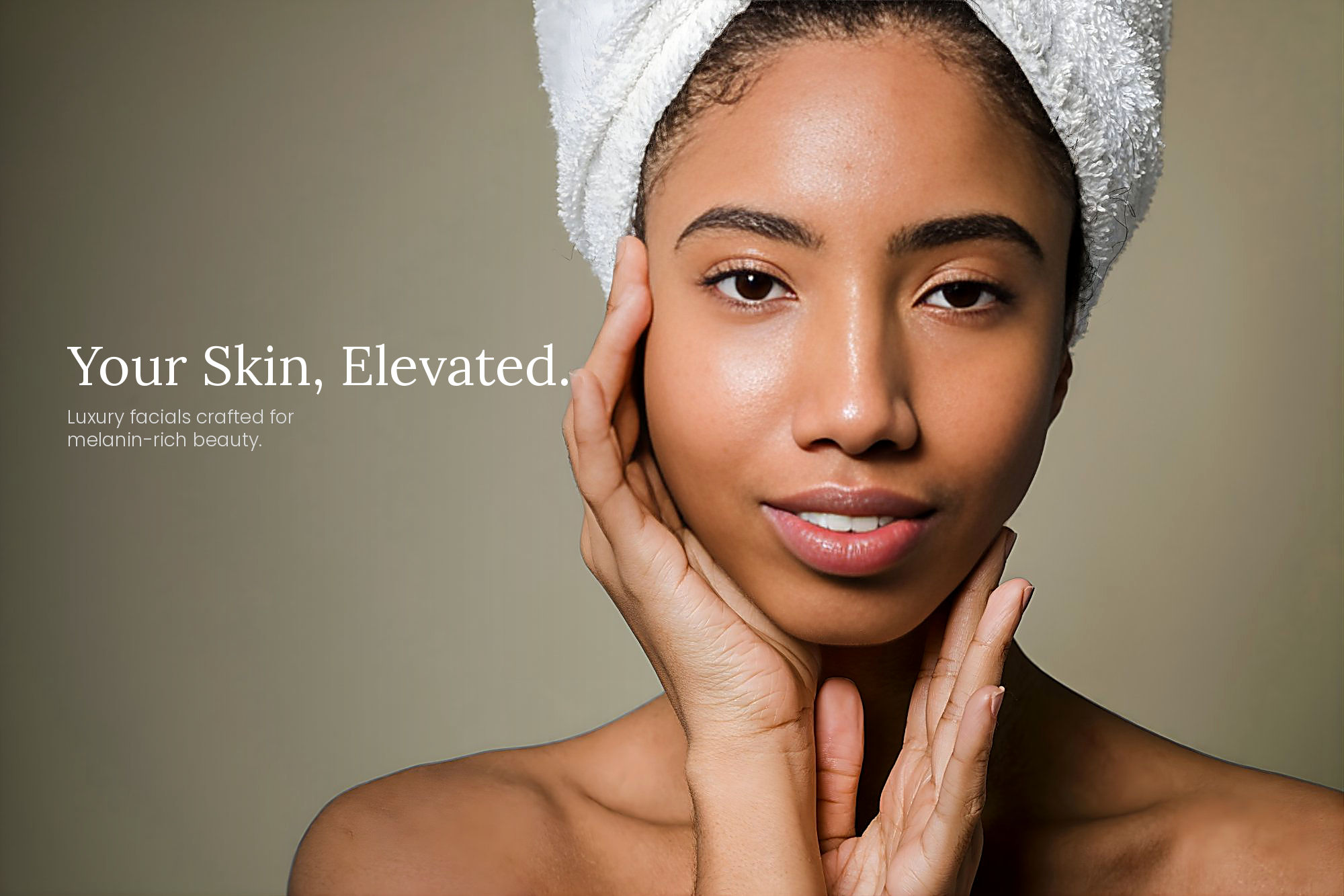 A woman with a towel wrapped around her head and clear, glowing skin touches her face with both hands, smiling softly.