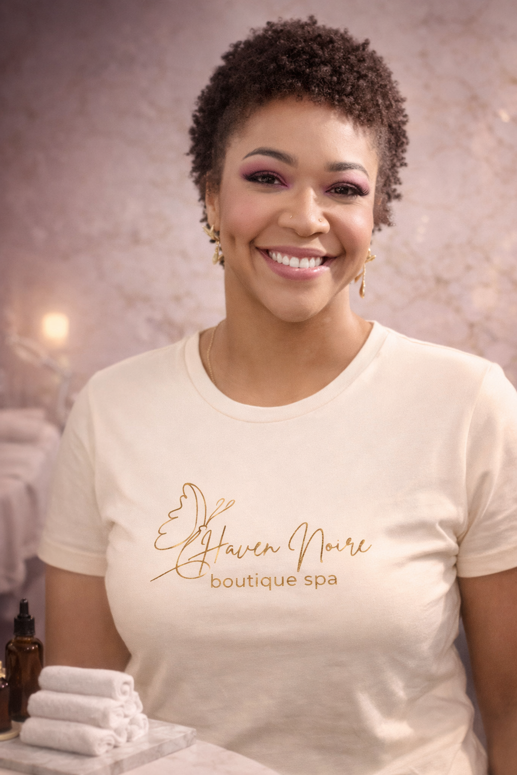 A woman with short, curly hair smiling and wearing a cream-colored t-shirt with the logo 'Haven Moire boutique spa,' standing in a spa or boutique setting.