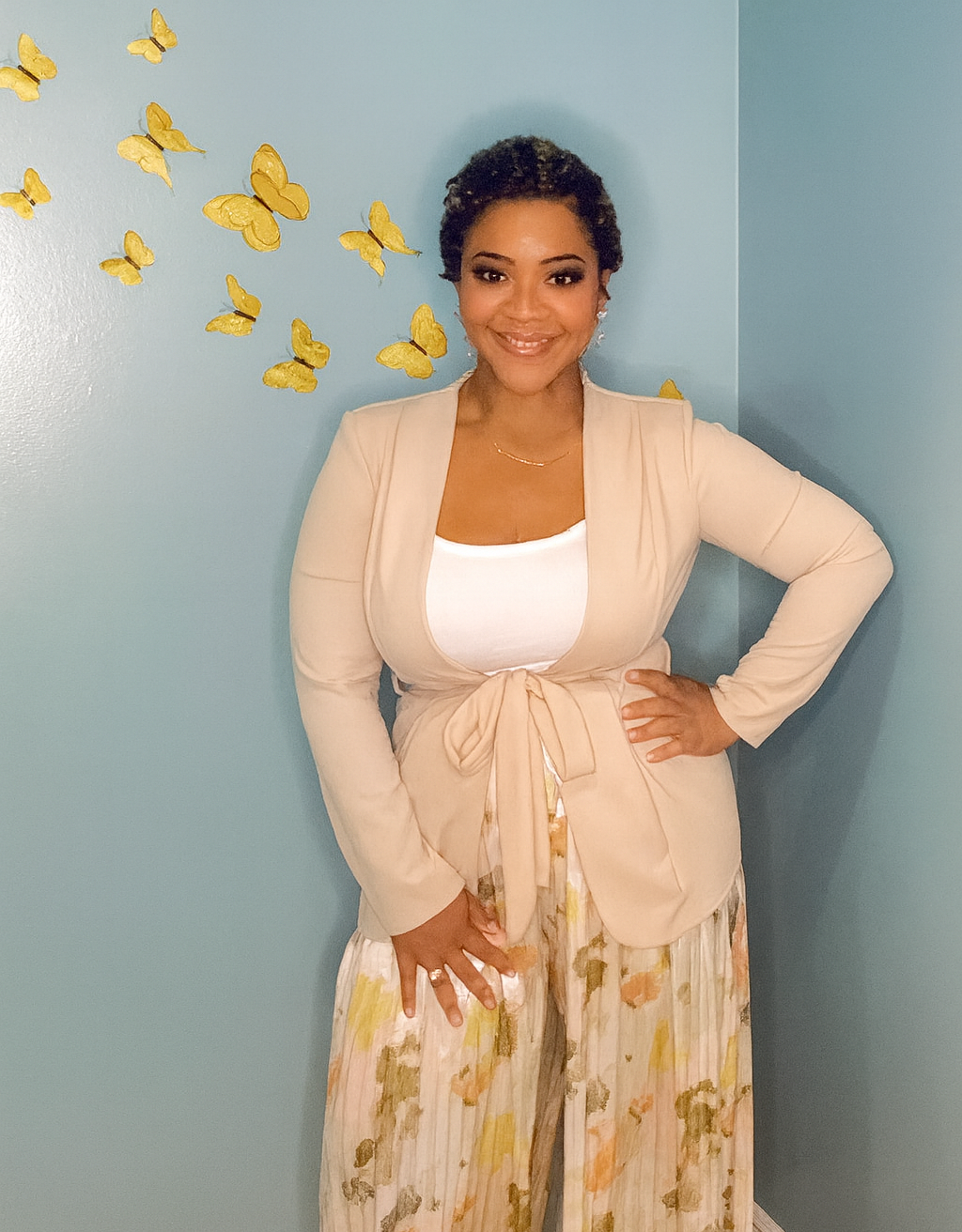 A woman standing against a blue wall with yellow butterfly wall decorations, wearing a beige blazer over a white top and floral-patterned pants, smiling with hand on hip.