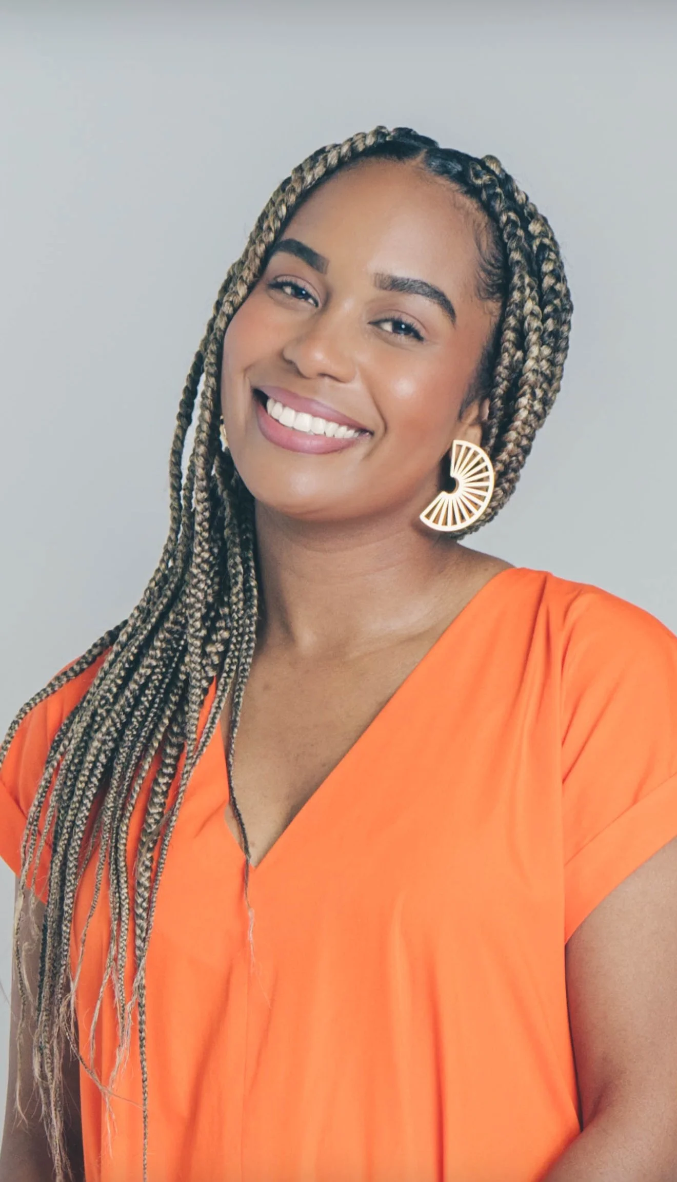 A woman with long braided hair, wearing a bright orange top and large, half-circular earrings, smiling at the camera against a plain background.