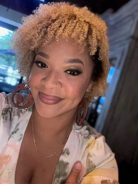 A woman with curly blonde hair and earrings smiling at the camera in a casual setting.