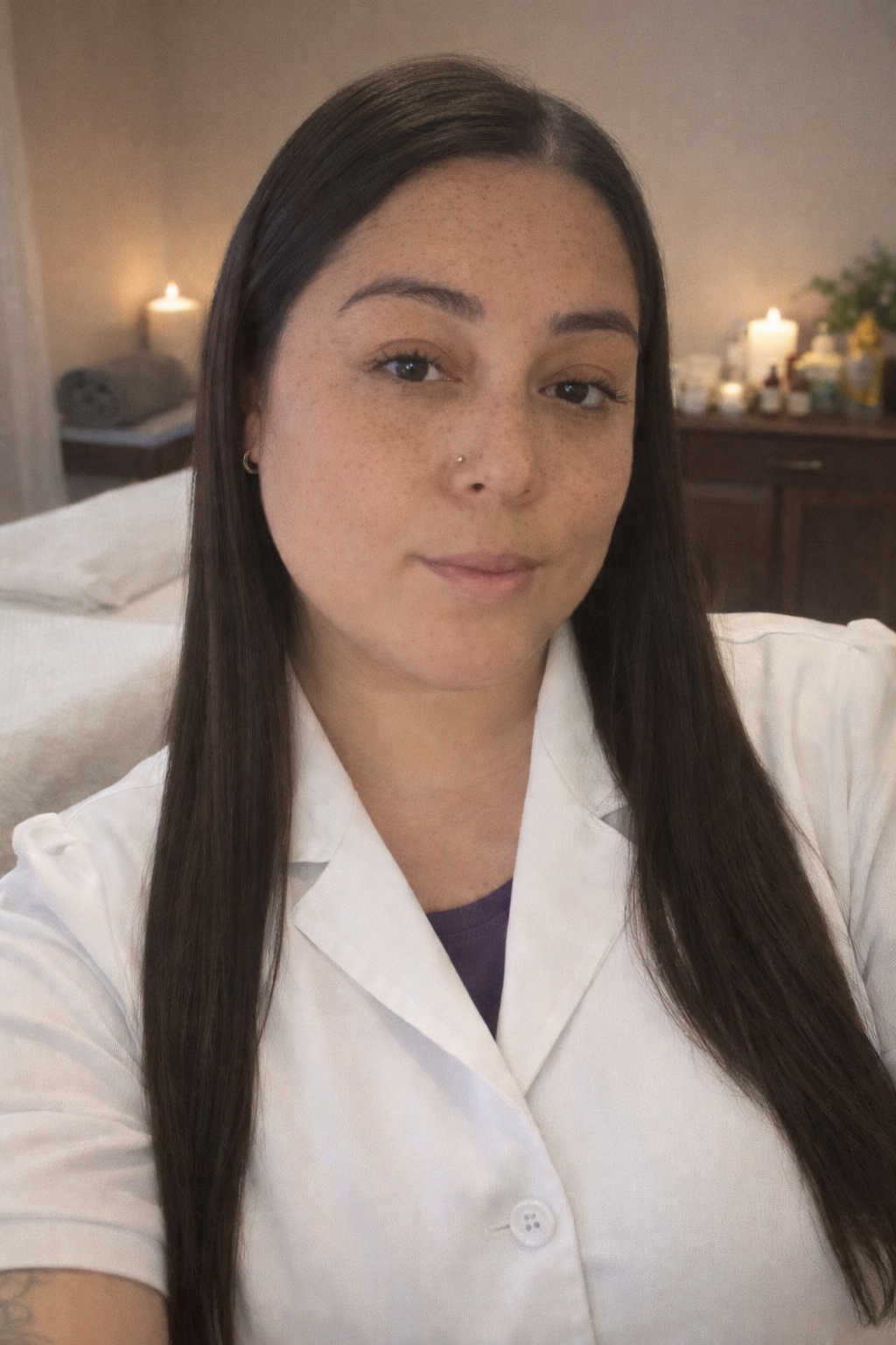 A woman with long dark hair, light skin with freckles, wearing a white button-up shirt, taking a selfie in a cozy room with candles and skincare products in the background.