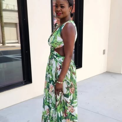A woman standing outside near a building, wearing a green and white floral dress with a sleeveless design, holding a small purse, and smiling.