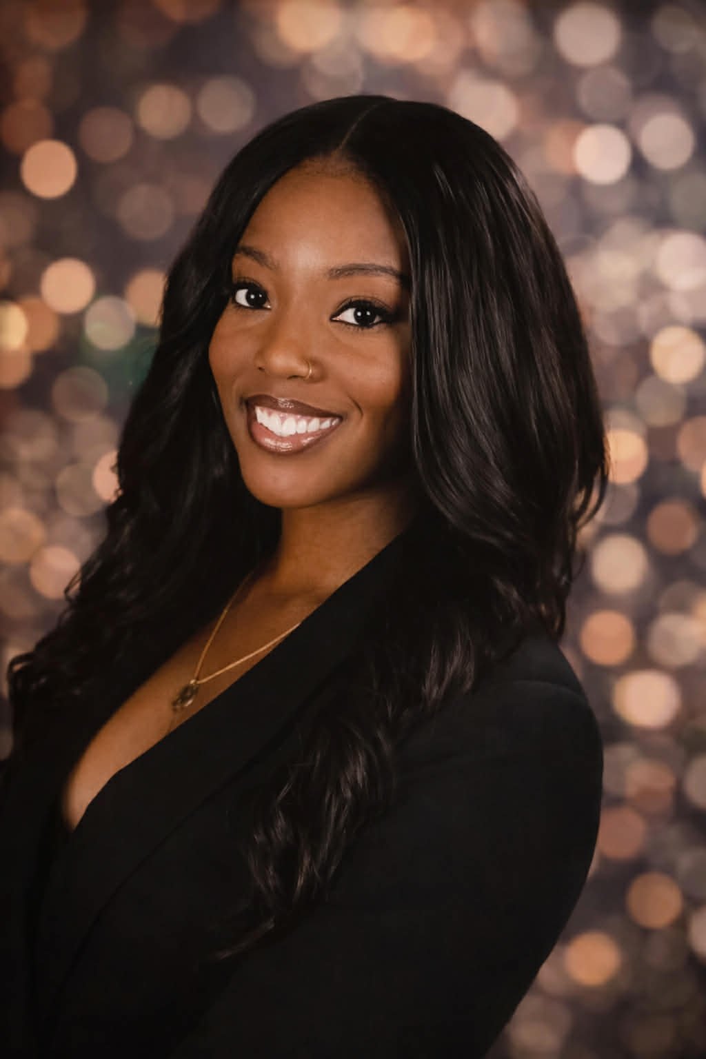 A smiling woman with long, wavy black hair, wearing a black blazer and a gold necklace, standing against a blurred background of lights.