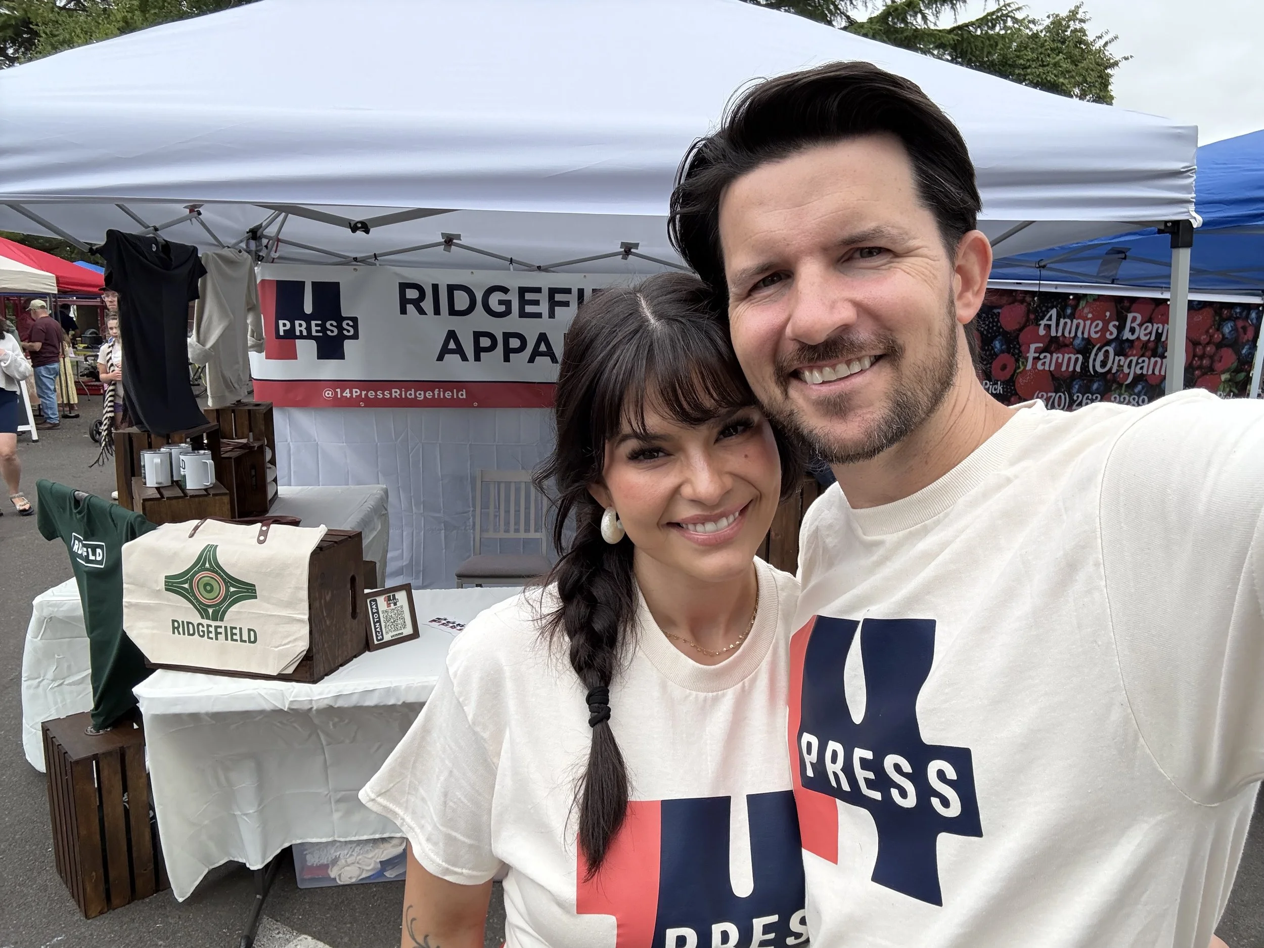Two people taking a selfie at an outdoor event, standing in front of a booth with