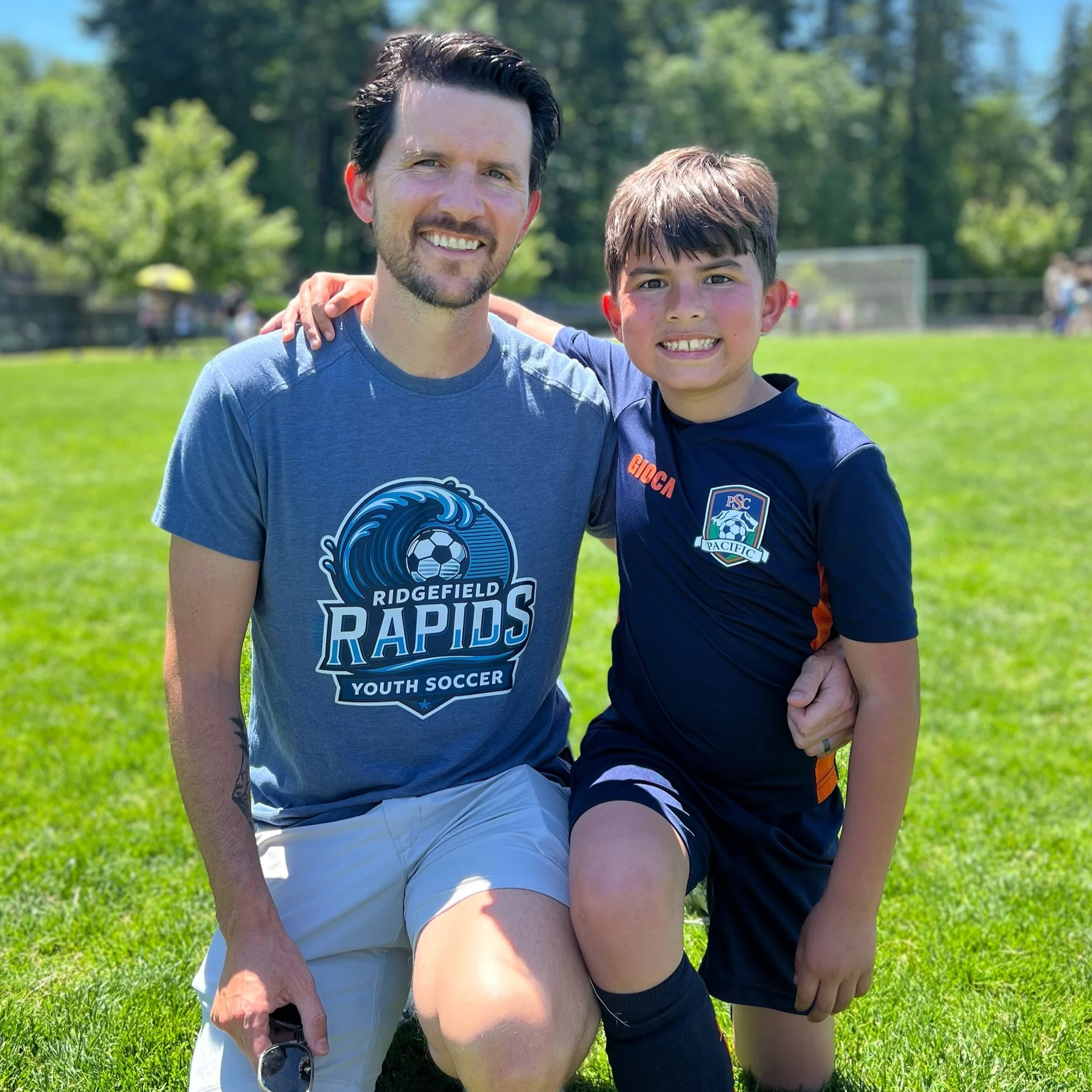 A man and a boy smiling and posing together on a grassy field on a sunny day. Both are wearing sports attire, with the man in a gray Ridgefield Rapids Youth Soccer T-shirt and the boy in a dark blue soccer uniform. The man has dark hair and a beard, and the boy has short brown hair. The man has his arm around the boy, who is kneeling with one knee on the ground.